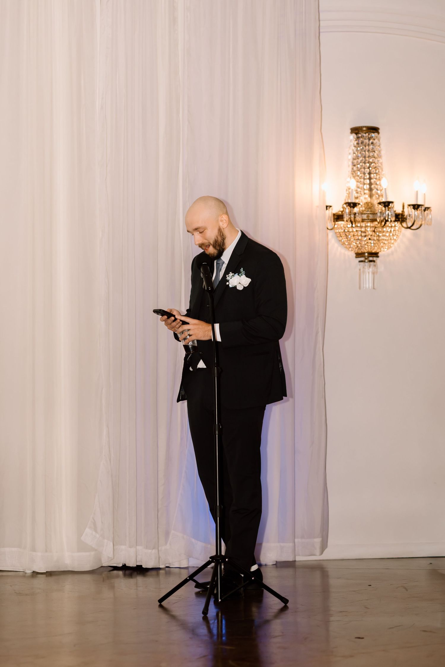 A person in a black tuxedo with a white boutonniere looks at their phone while standing near an elegant chandelier.