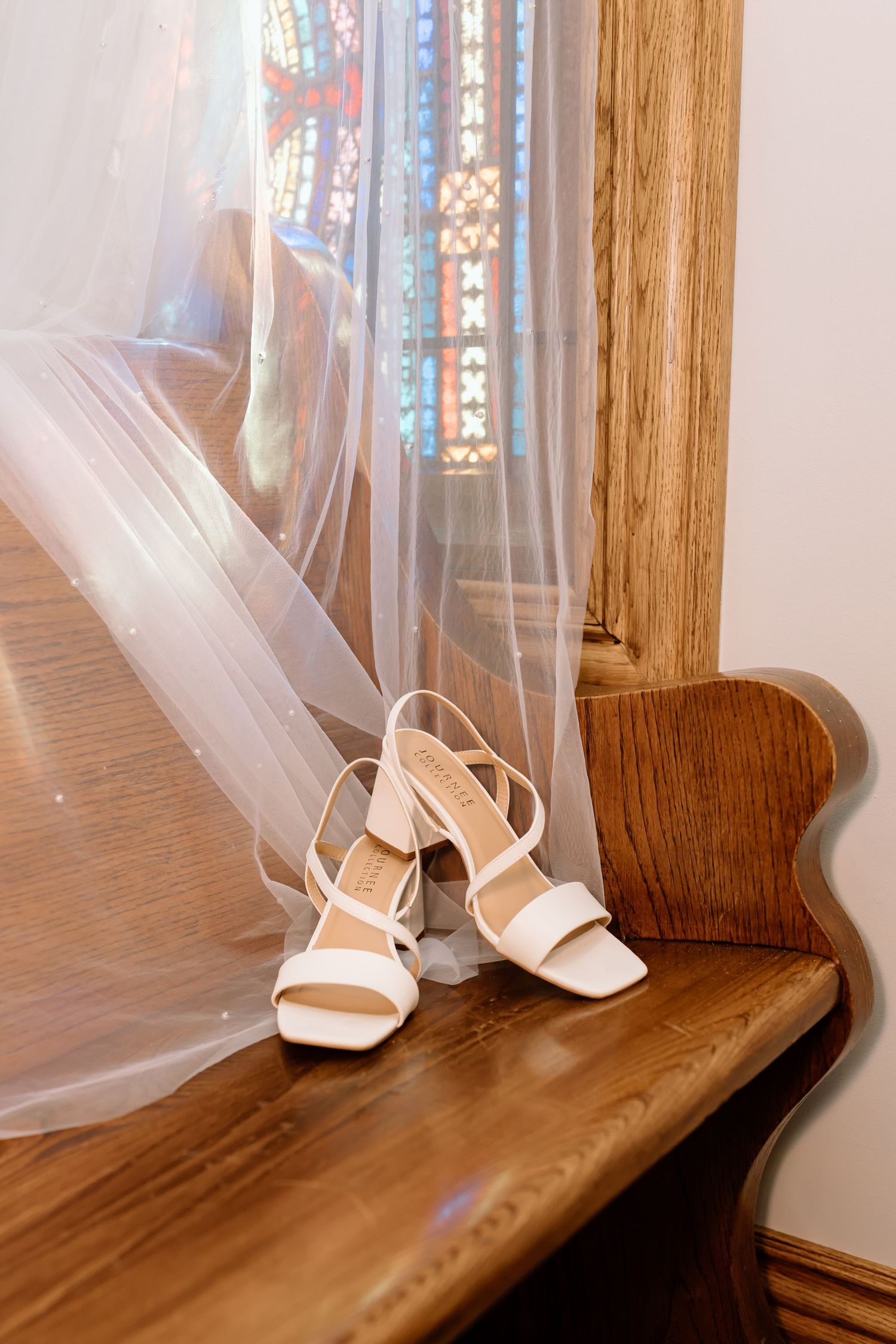 White wedding shoes sit on a wooden windowsill with sheer curtains and stained glass windows in the background.