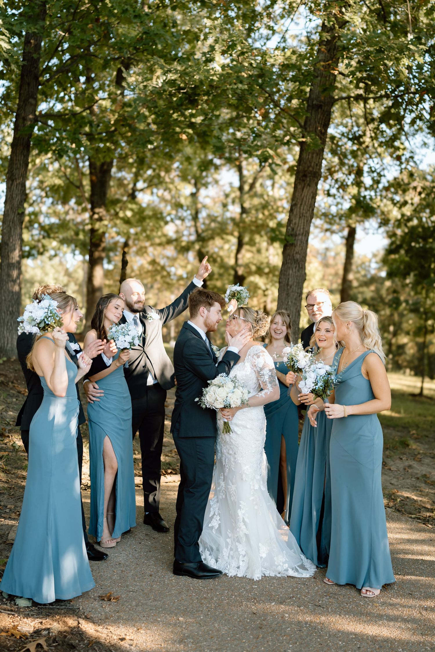 Wedding party gathers on a tree-lined path in matching light blue dresses and dark suits for outdoor celebration.