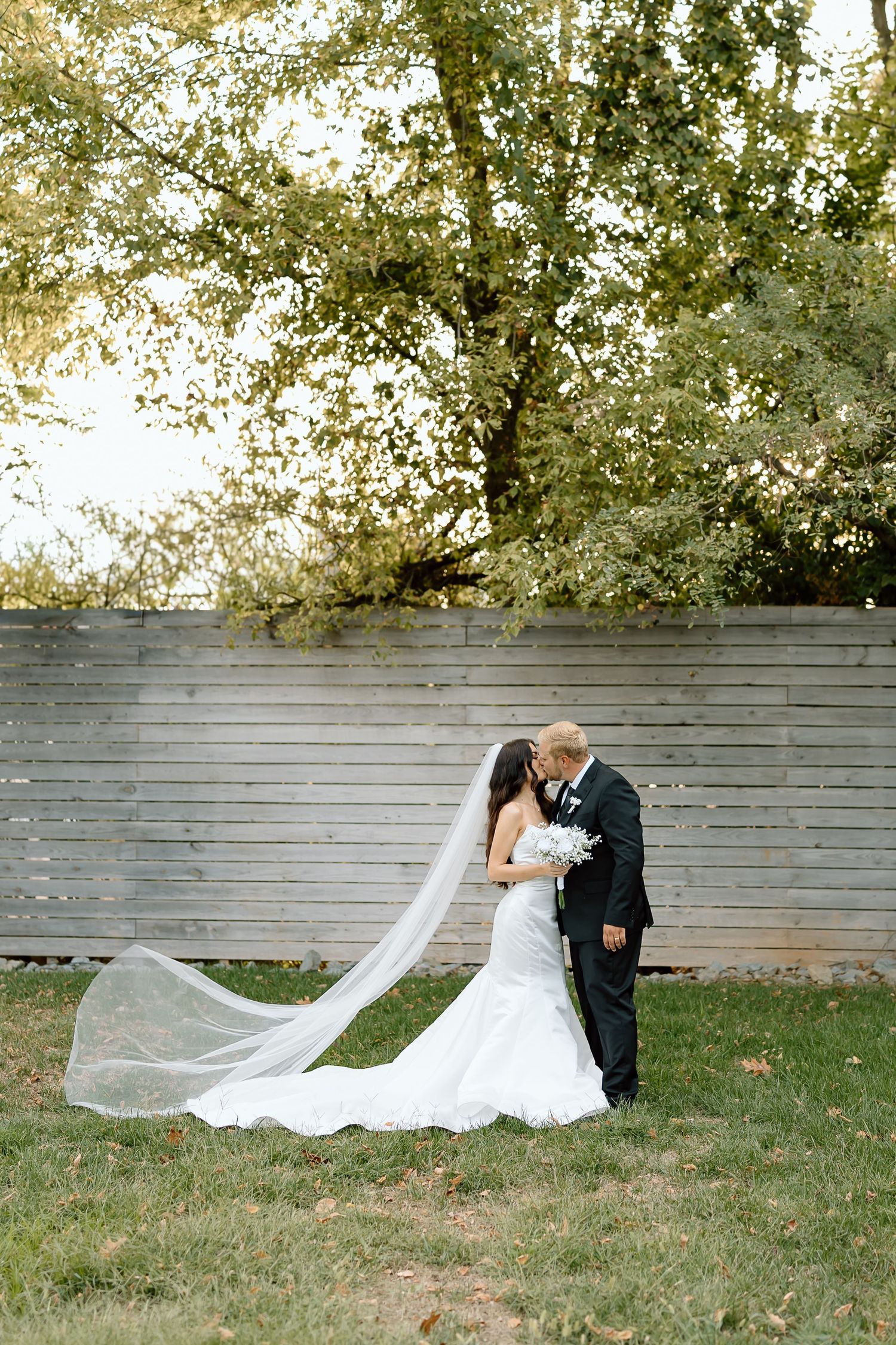 A couple shares a romantic moment in their wedding attire against a wooden fence backdrop with flowing veil and autumn trees.