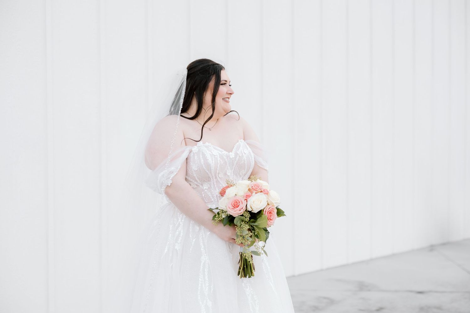 A bride in a flowing white dress holds a pink and white rose bouquet against a bright white backdrop.