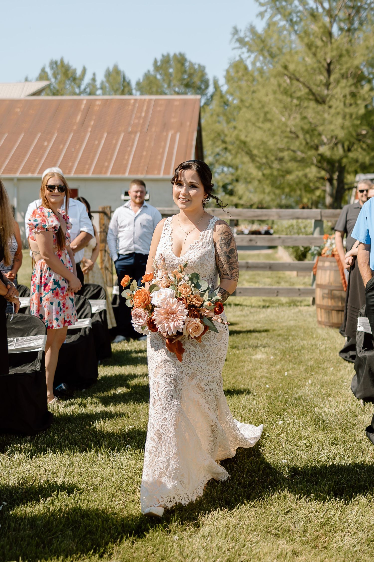 A bride in a lace wedding dress holds a peach and coral bouquet at a rustic barn venue on a sunny day.