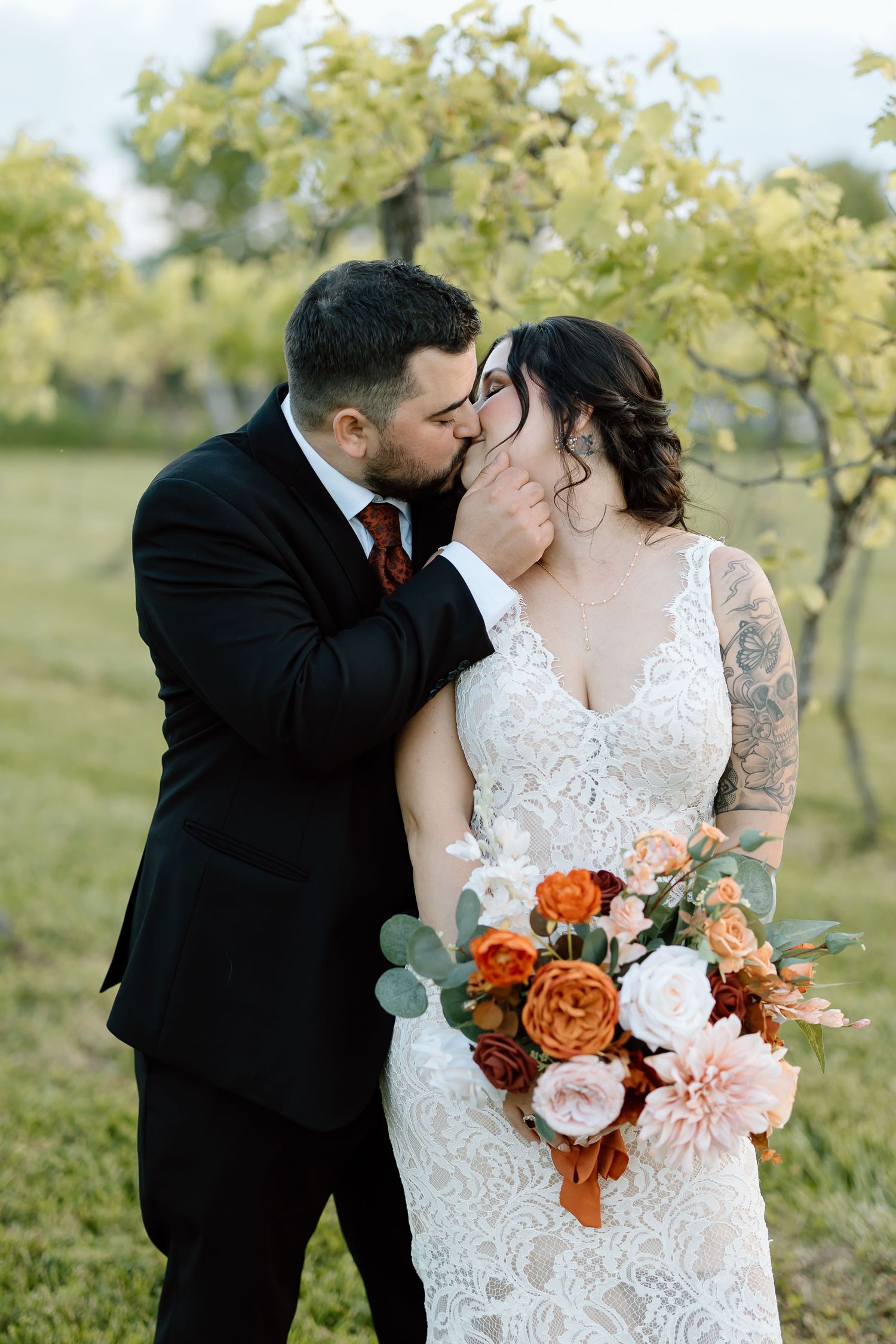Romantic couple shares tender kiss in garden setting with lush orange and pink wedding bouquet in lace dress and suit.