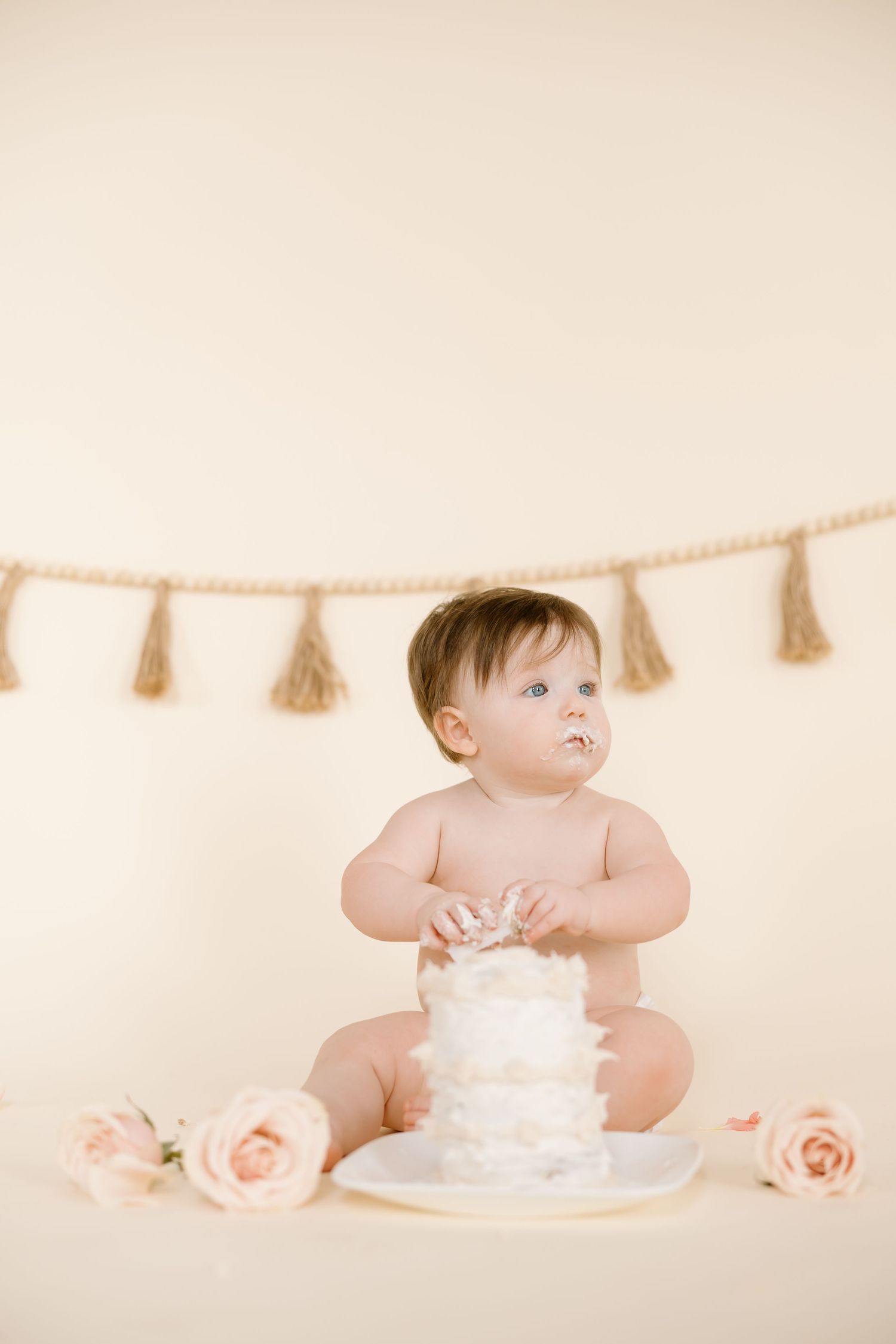 Small child in diaper reaching toward white cake with pink roses and tassel garland backdrop.