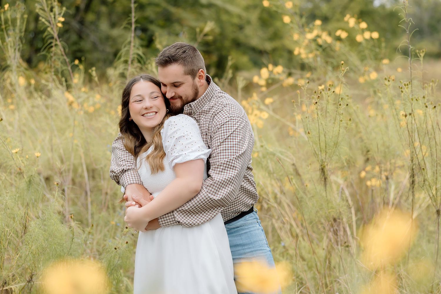 A couple shares a warm embrace while standing among yellow wildflowers in a sunny meadow.