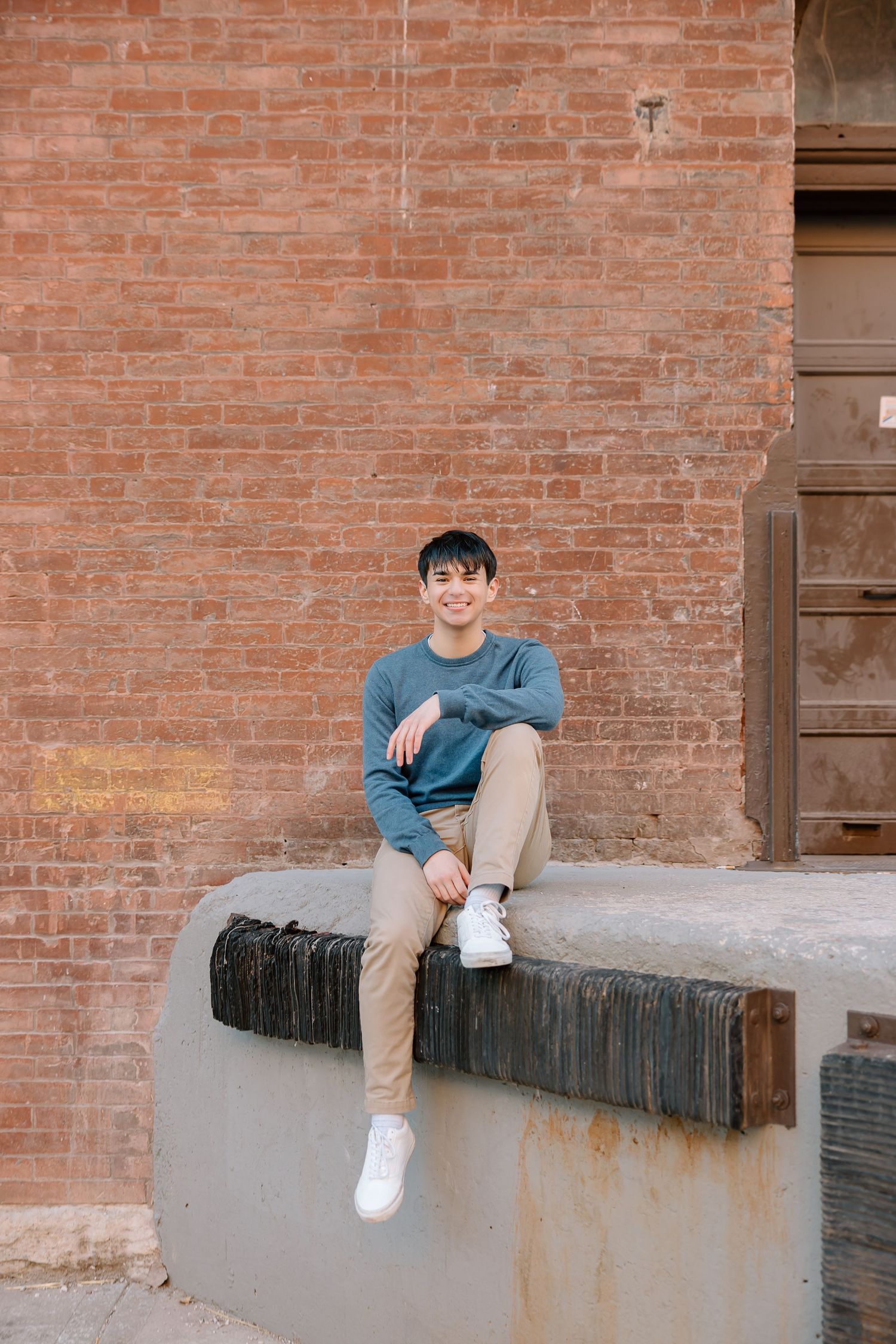 A casual outdoor portrait against a rustic brick wall, sitting on a concrete ledge in a blue sweater and khaki pants.