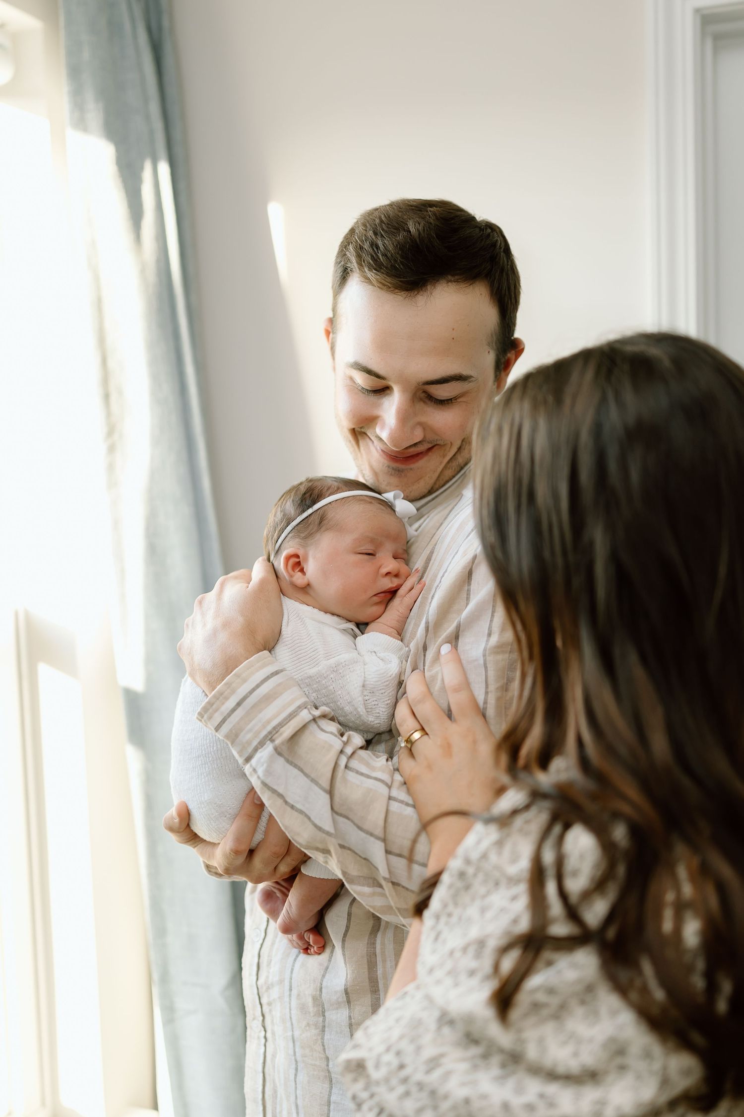 A tender family moment as parents share loving kisses with their baby near a bright window.