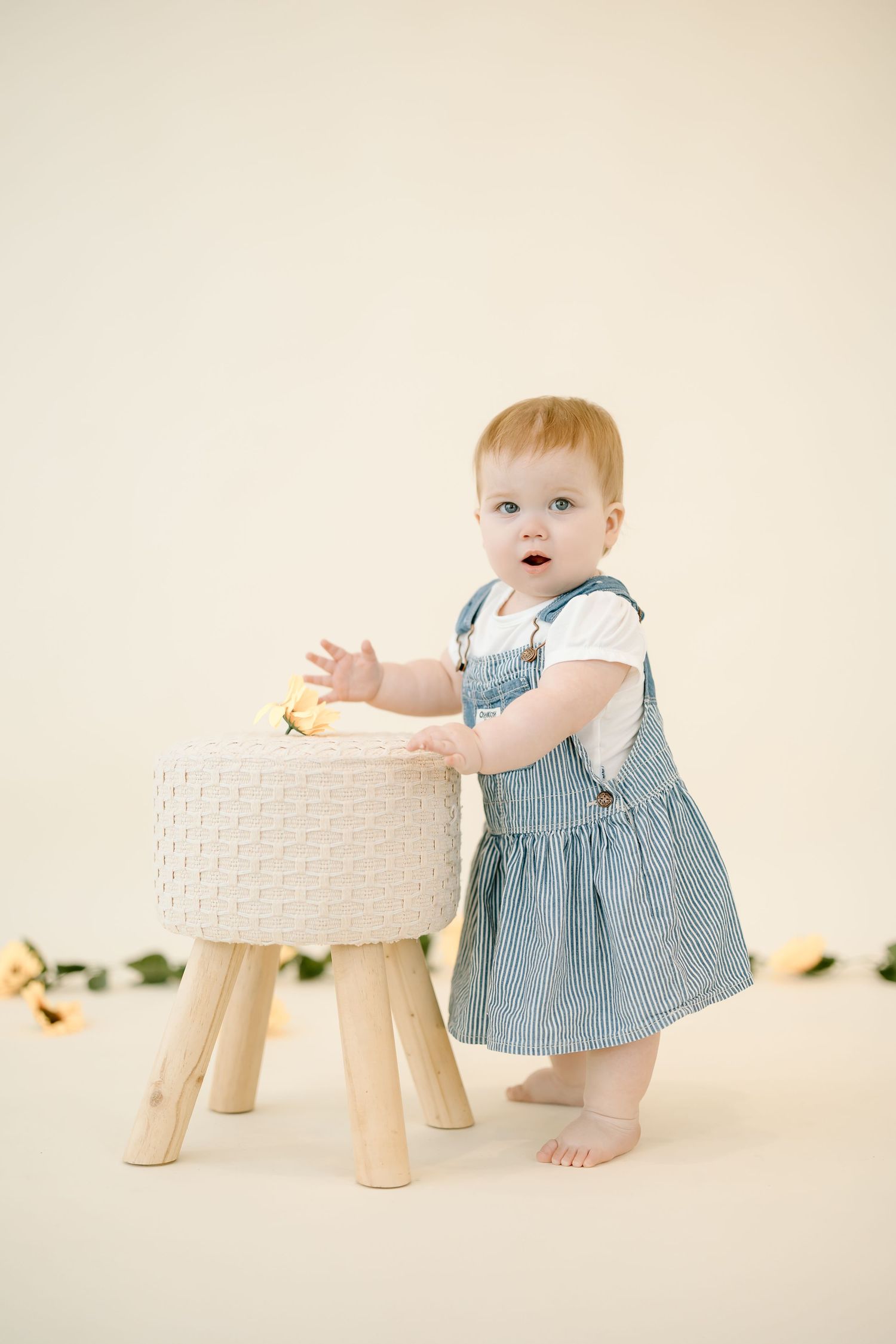 A small child in a blue dress stands beside a white woven stool against a cream colored backdrop.