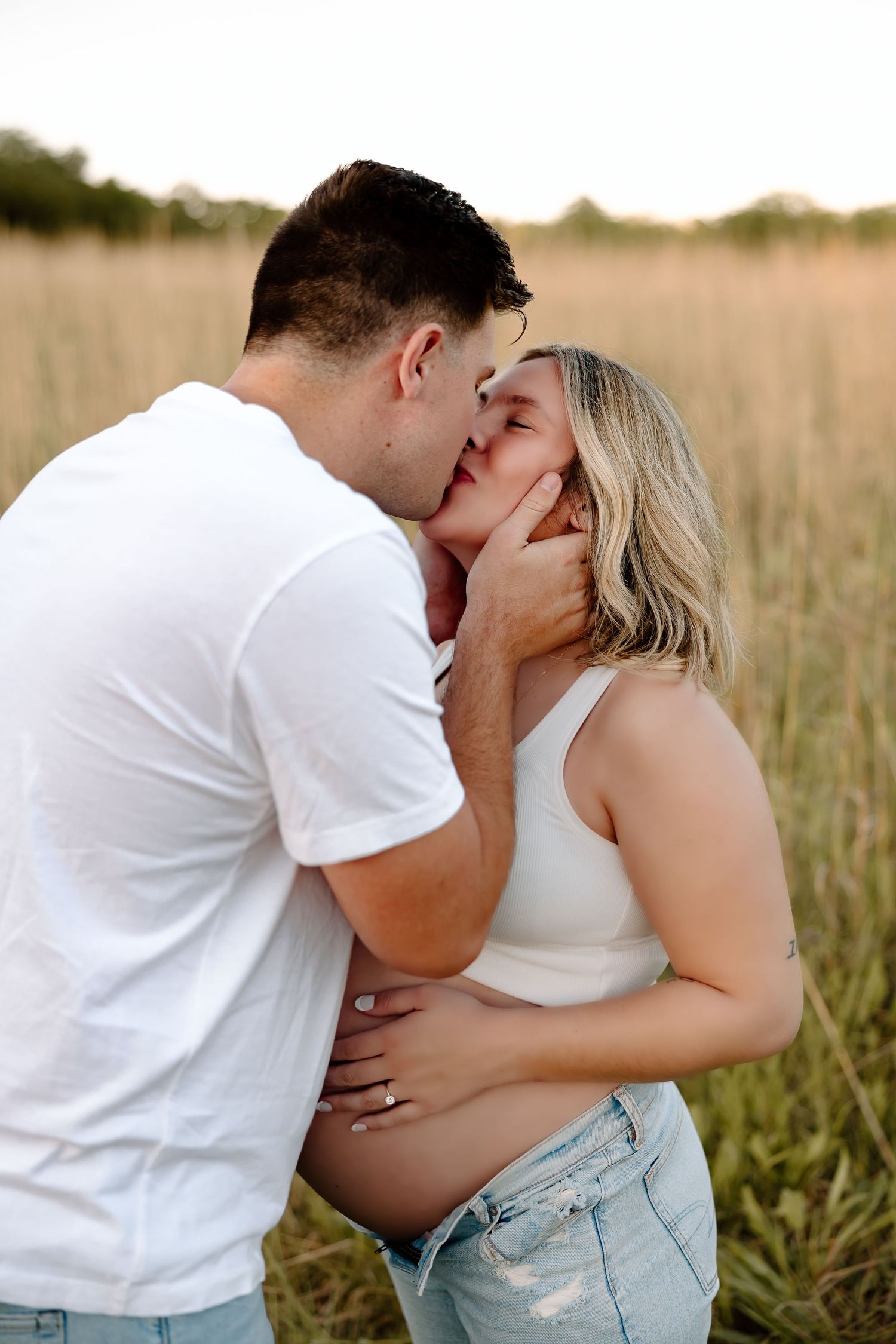 A romantic couple in white shirts share tender kisses in a sunlit field at golden hour.