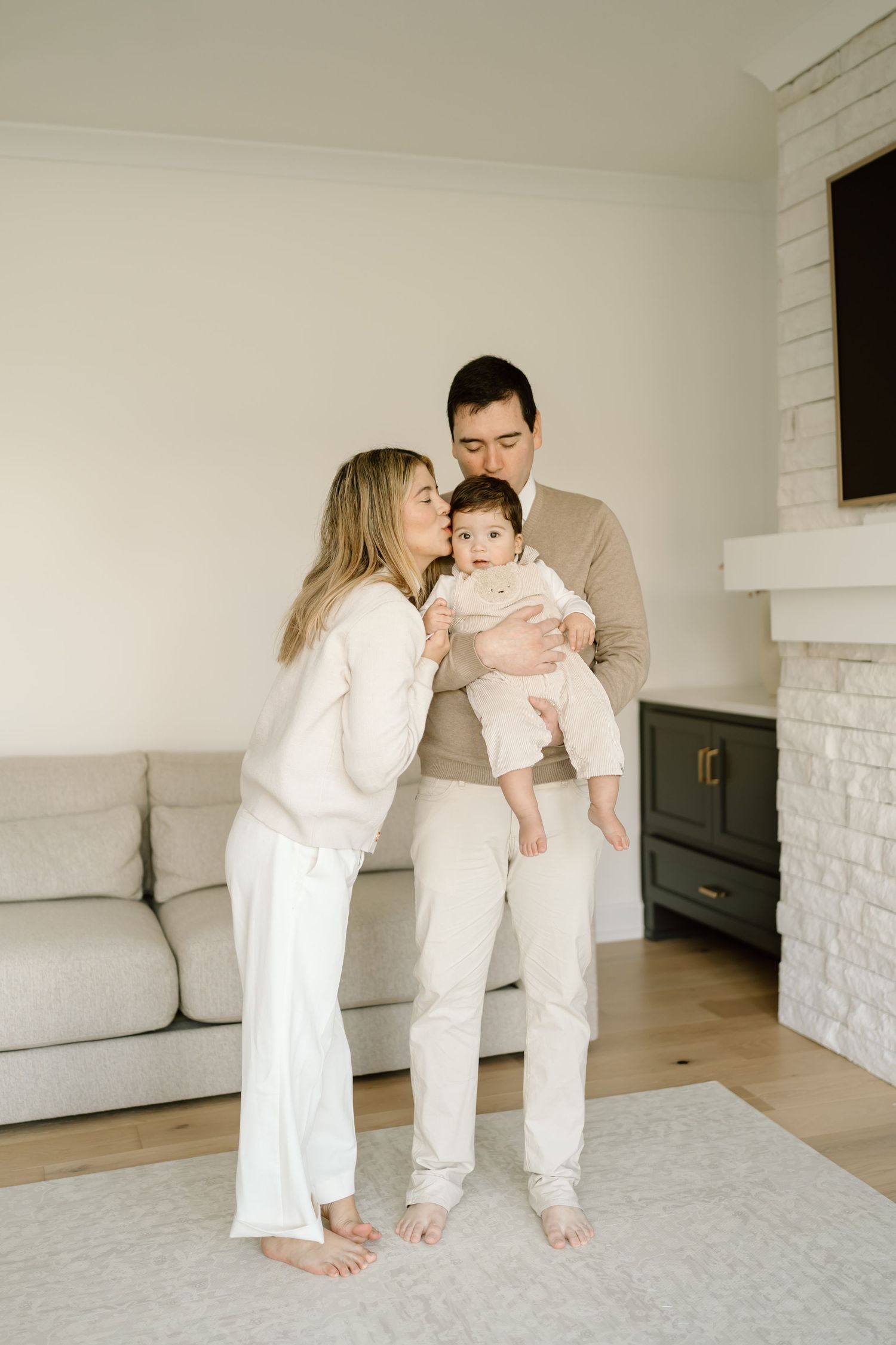 A family in white clothing shares tender moments while sitting on a light carpet in a minimalist room.