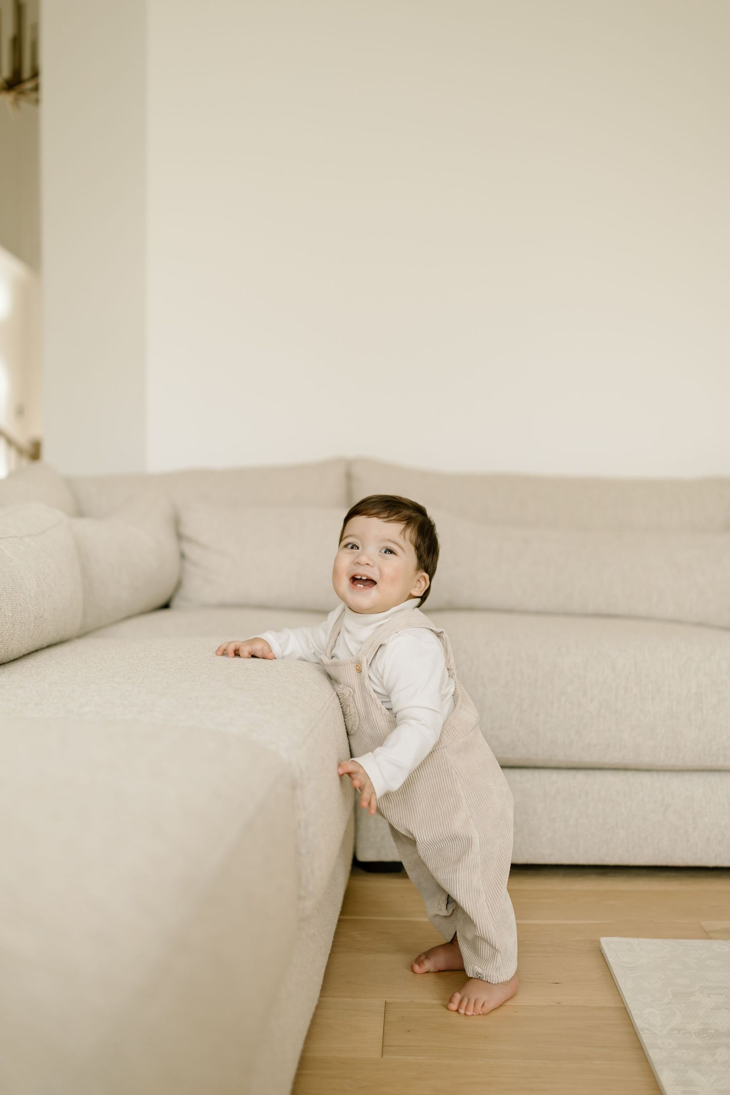 A small child in a beige romper and white shirt stands barefoot while holding onto a light-colored sofa.