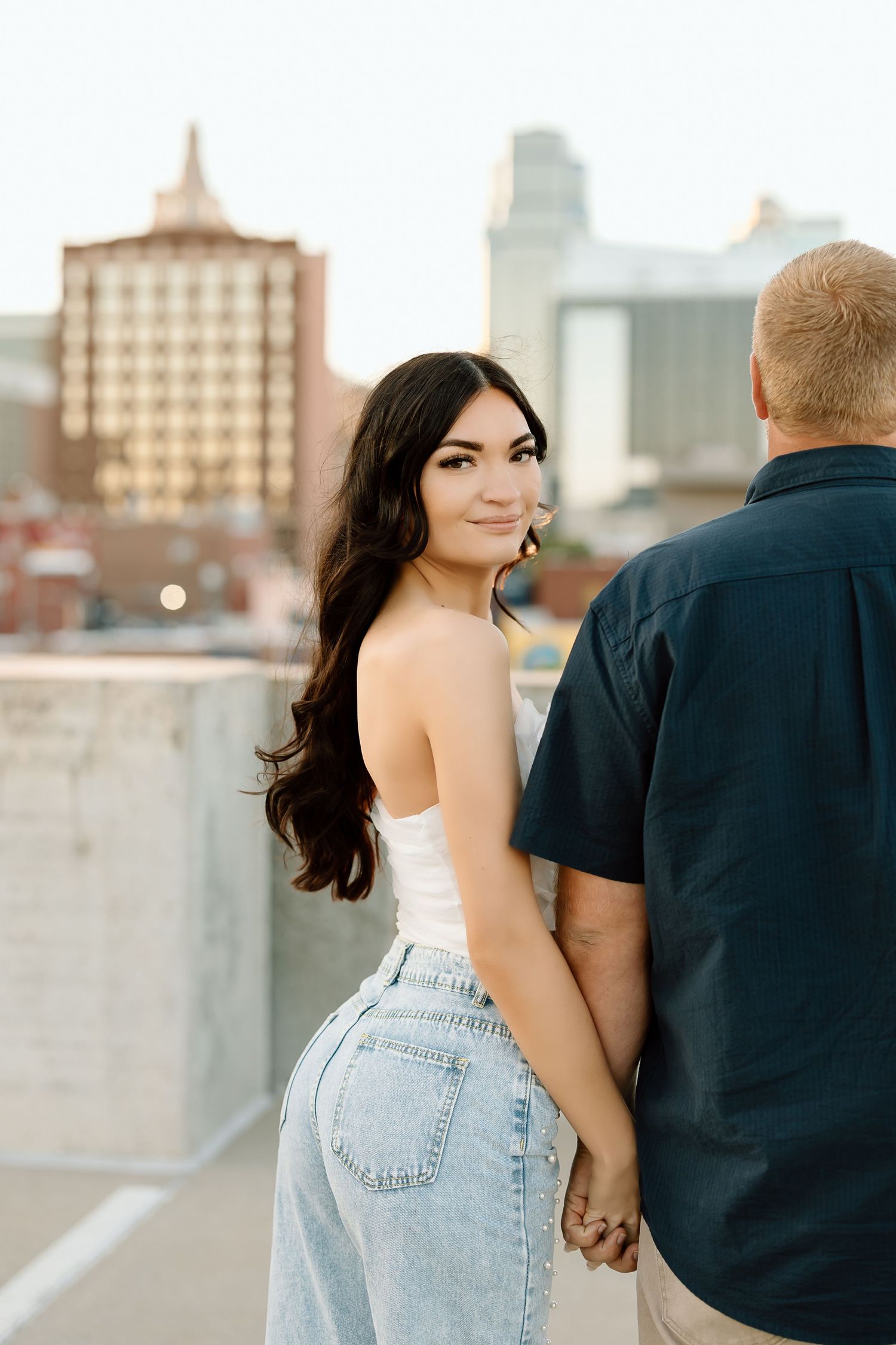 A couple poses together on a rooftop with an urban cityscape backdrop during an engagement photoshoot.