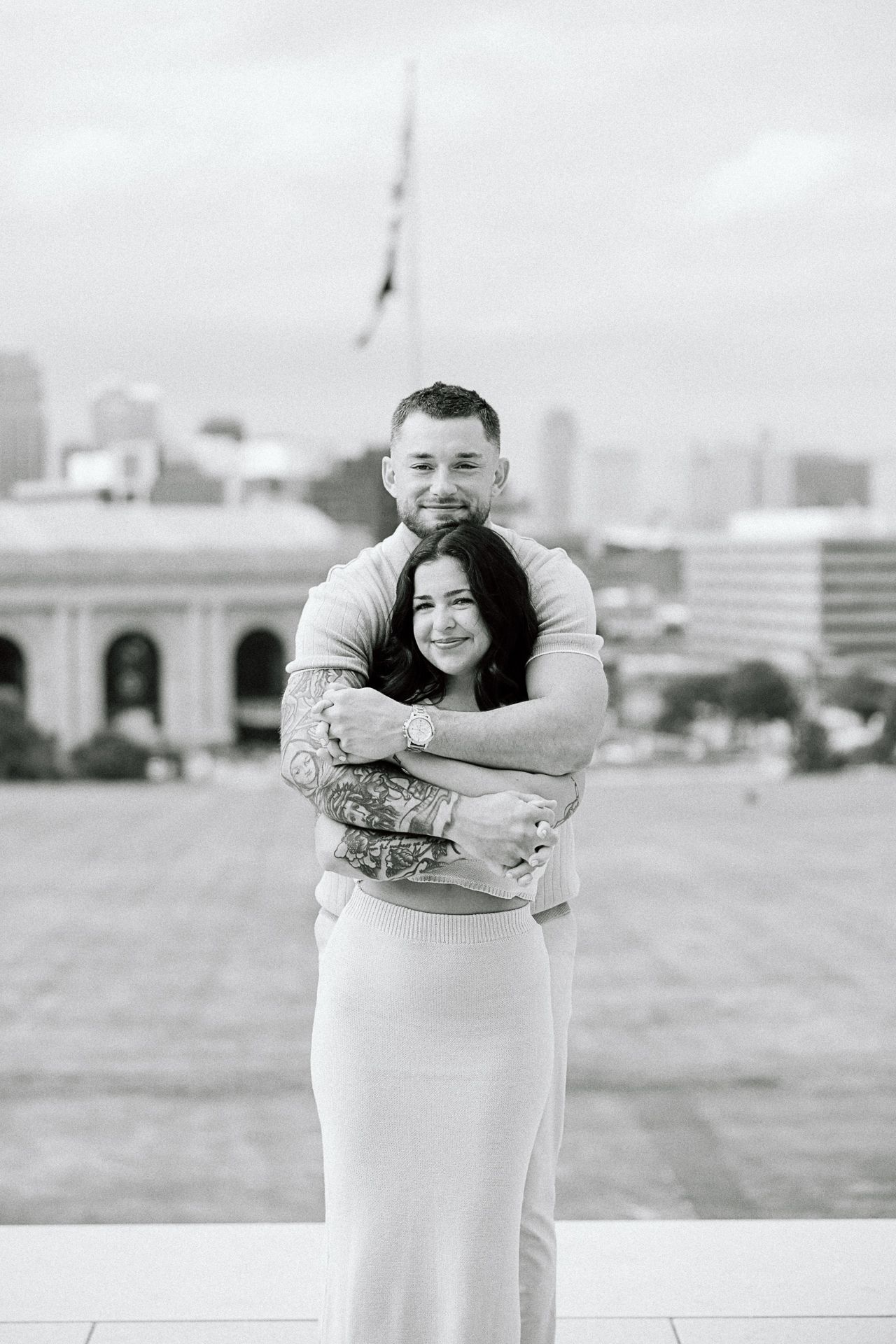 A couple hugs and laughs together during their outdoor portrait session with city buildings in the background.