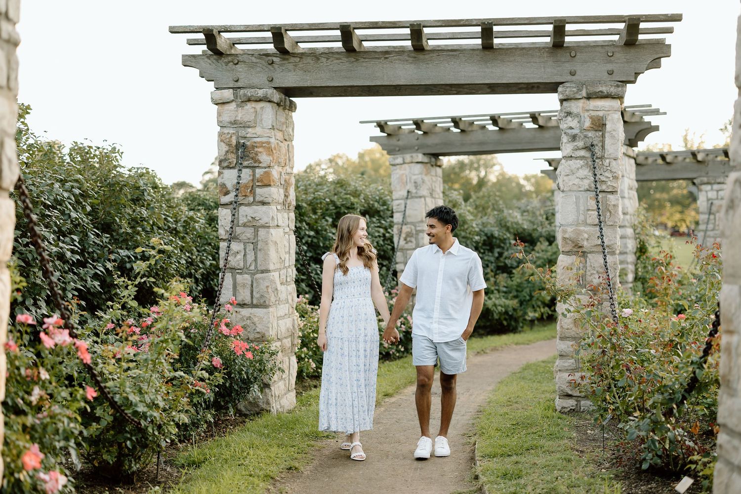 A couple walks through a romantic garden pergola surrounded by blooming roses and stone columns on a sunny day.
