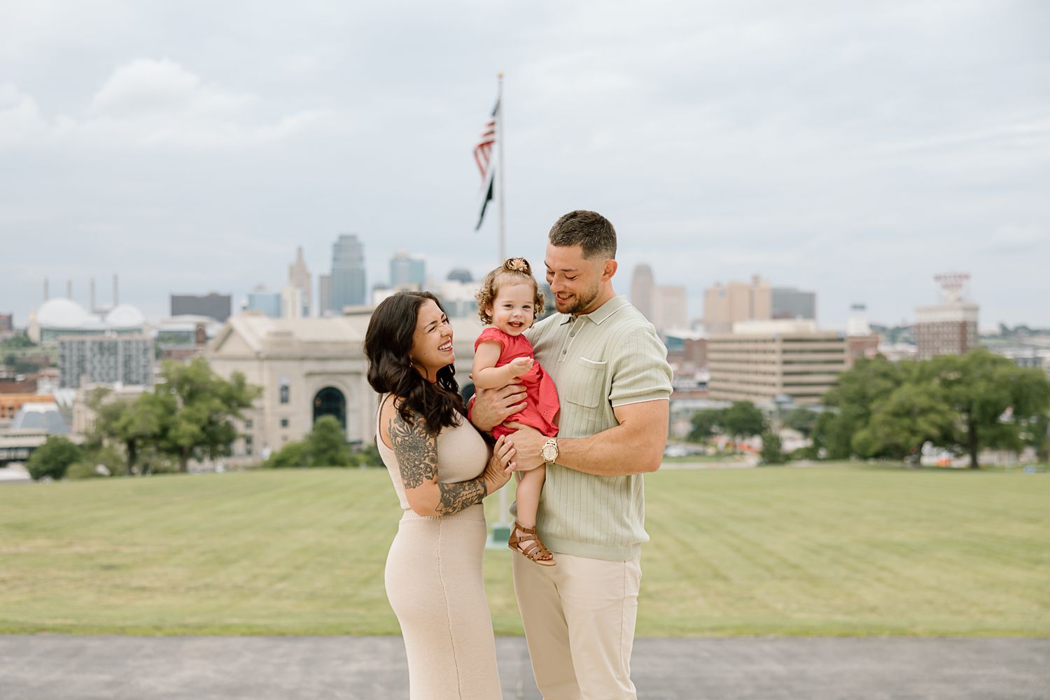 A family stands together on a grassy hill overlooking the Kansas City skyline with an American flag in the background.