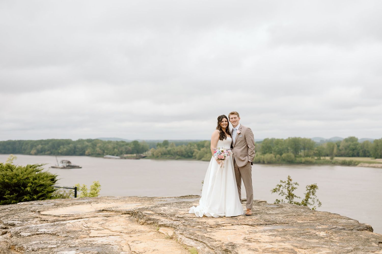 A couple in wedding attire stands together on a scenic stone overlook with a lake and cloudy sky in the background.