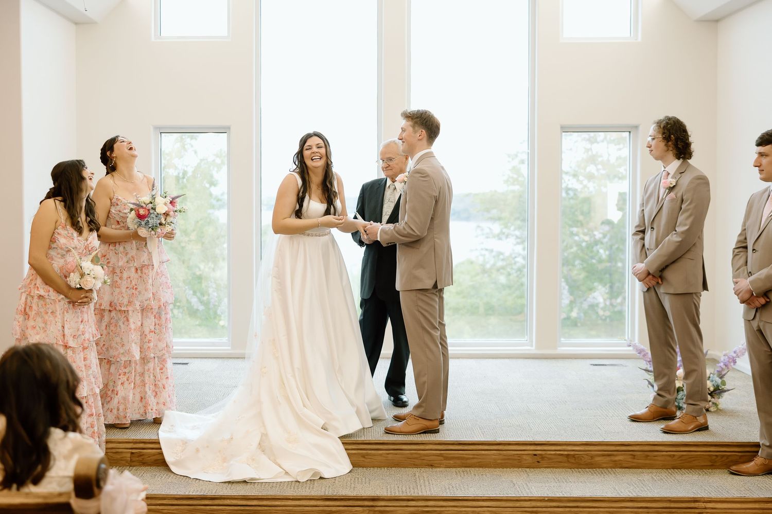 A wedding ceremony takes place in a bright sunlit room with large windows and a wedding party standing on a wooden platform.