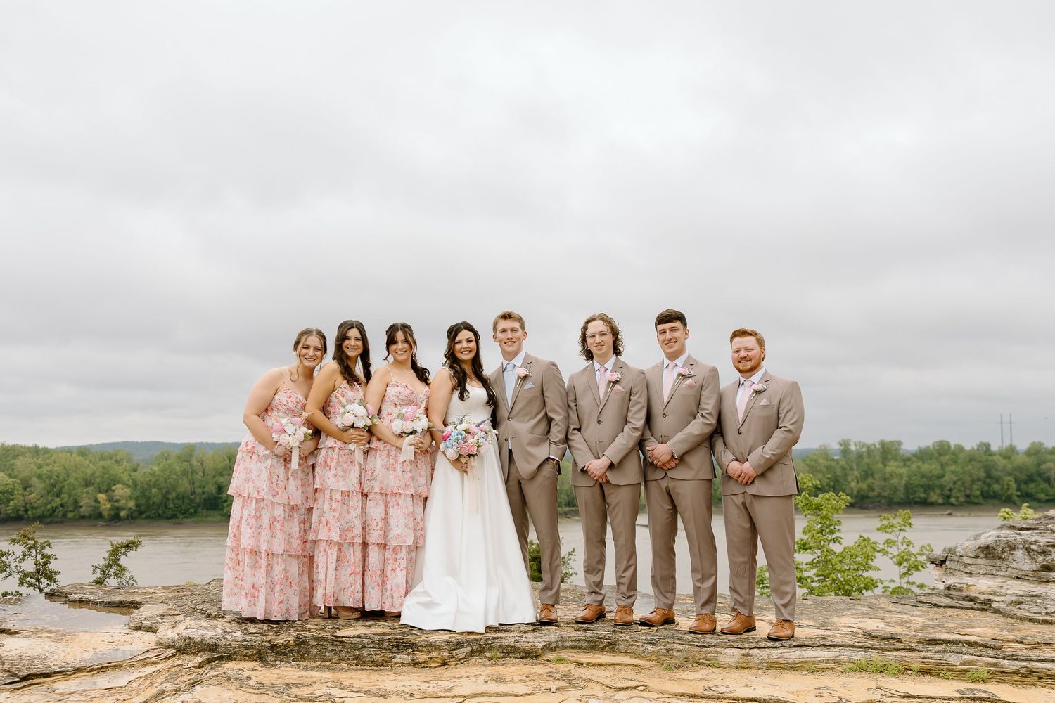 Wedding party in pink dresses and tan suits poses together on a rocky beach landscape under cloudy skies.