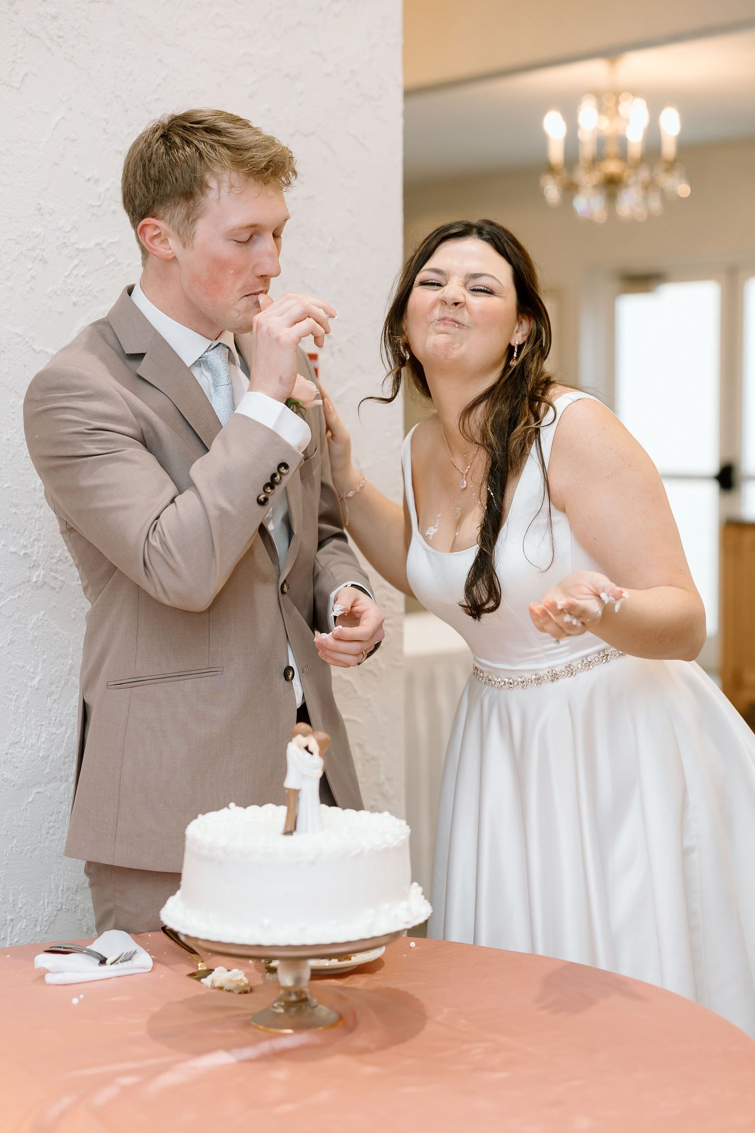 Wedding couple shares playful cake feeding moments during reception at elegant venue with chandelier lighting.