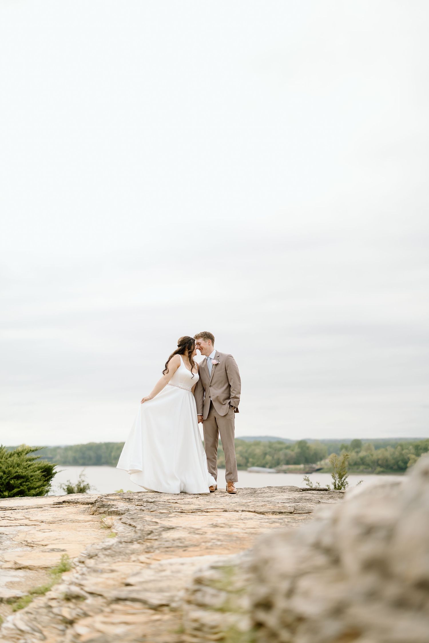 A couple in wedding attire shares an intimate moment on a scenic rocky cliff overlooking a lake and forest landscape.