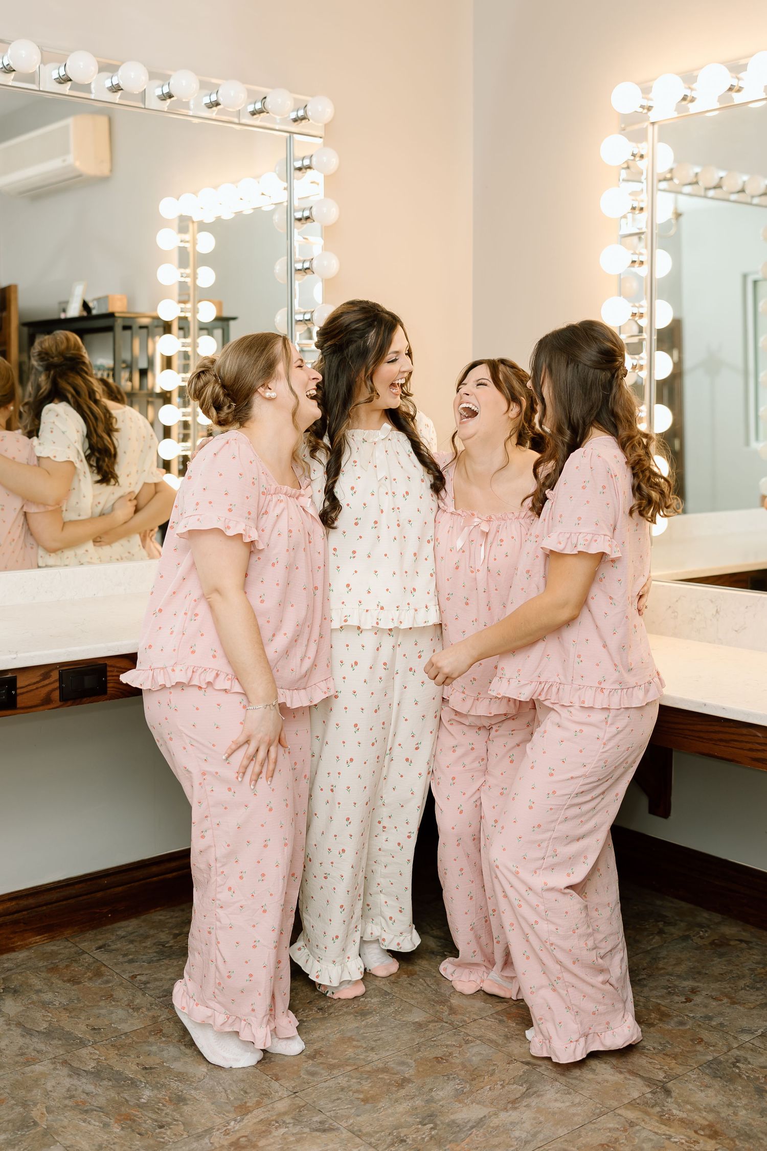 A group in matching pink pajamas and one in white share a joyful moment together in a dressing room with illuminated mirrors.