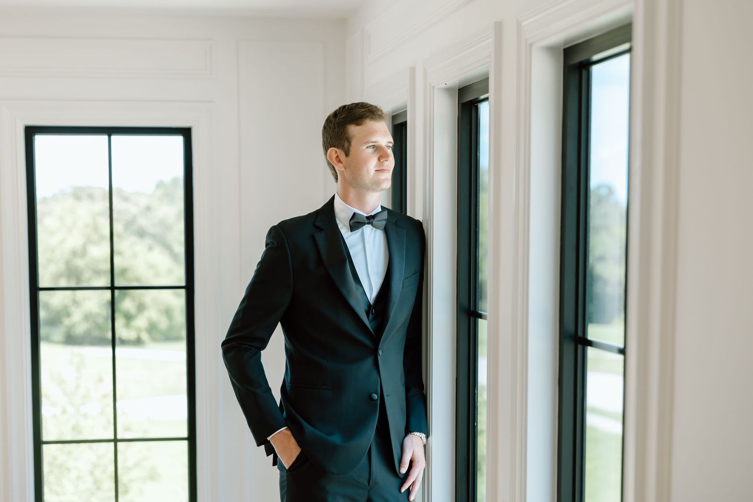 Groom in black tuxedo with bow tie standing by window, gazing outside at green landscape.