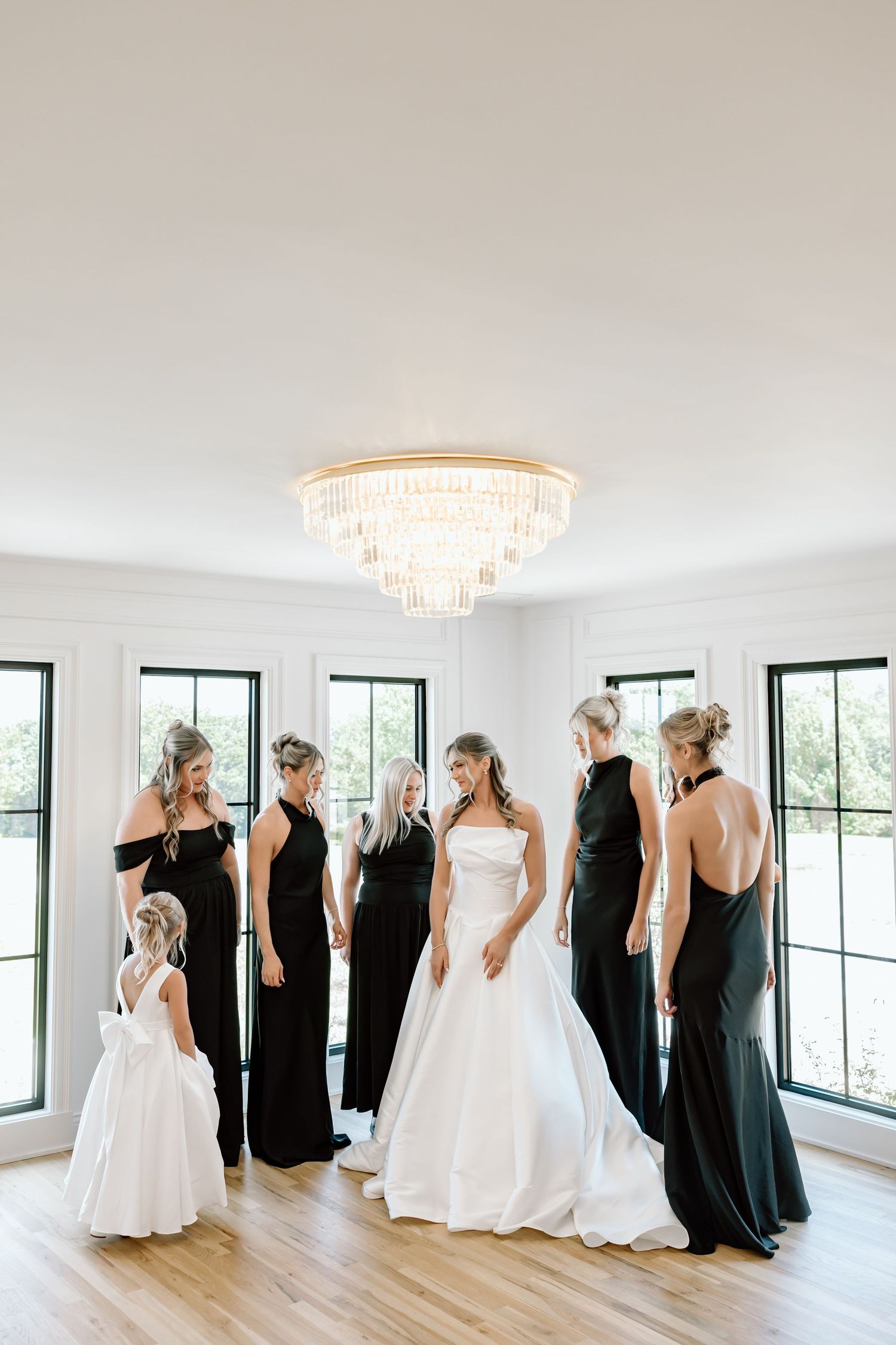 Bride in white gown with bridesmaids in black dresses and flower girl in white room with chandelier.