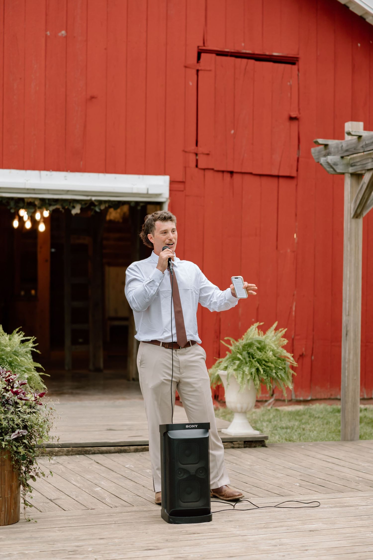 Man giving a speech outdoors on a wooden deck near a red barn, holding a microphone and drink at a wedding reception.