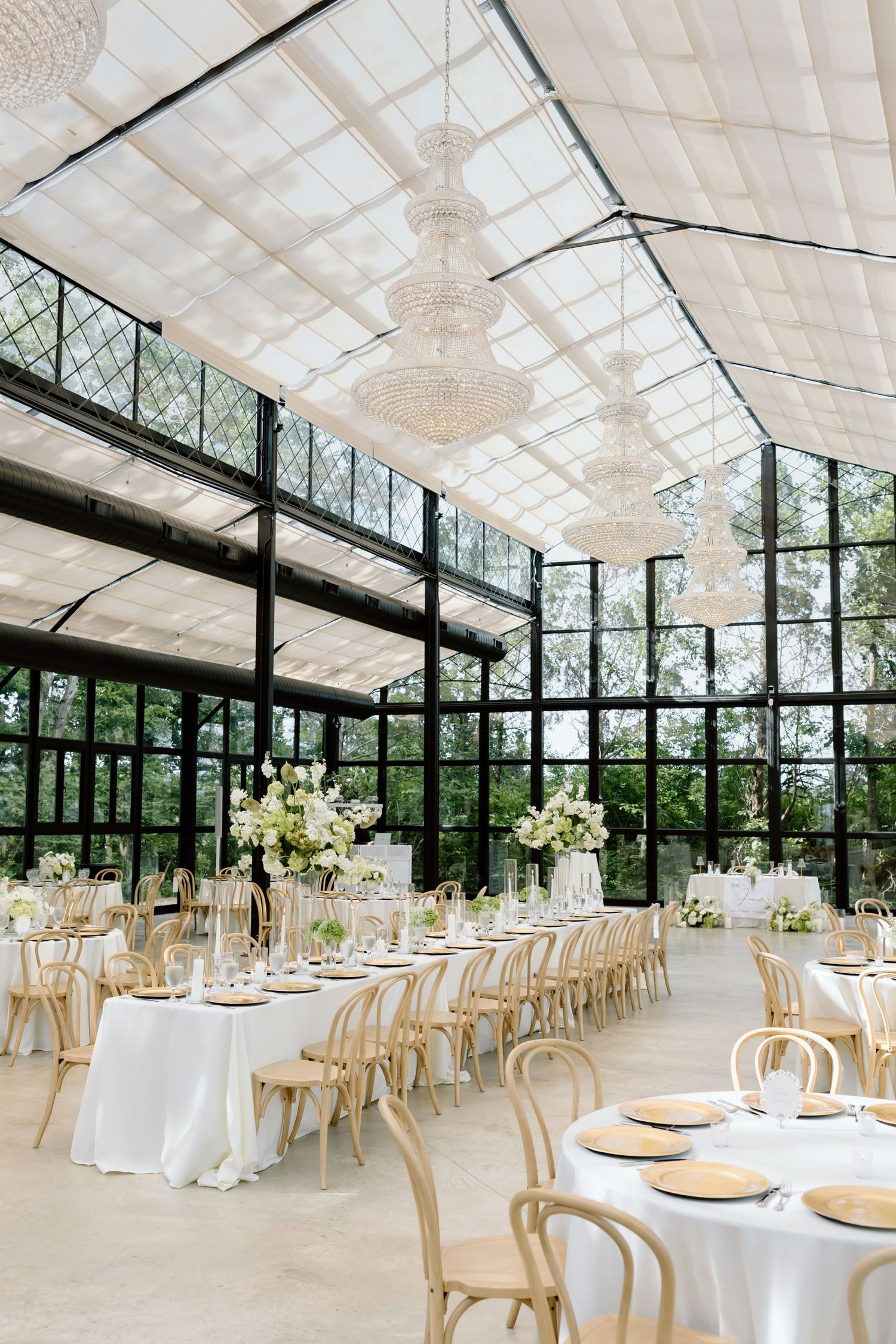 Elegant greenhouse wedding reception with crystal chandeliers, white floral centerpieces, and natural wood chairs under glass ceiling.