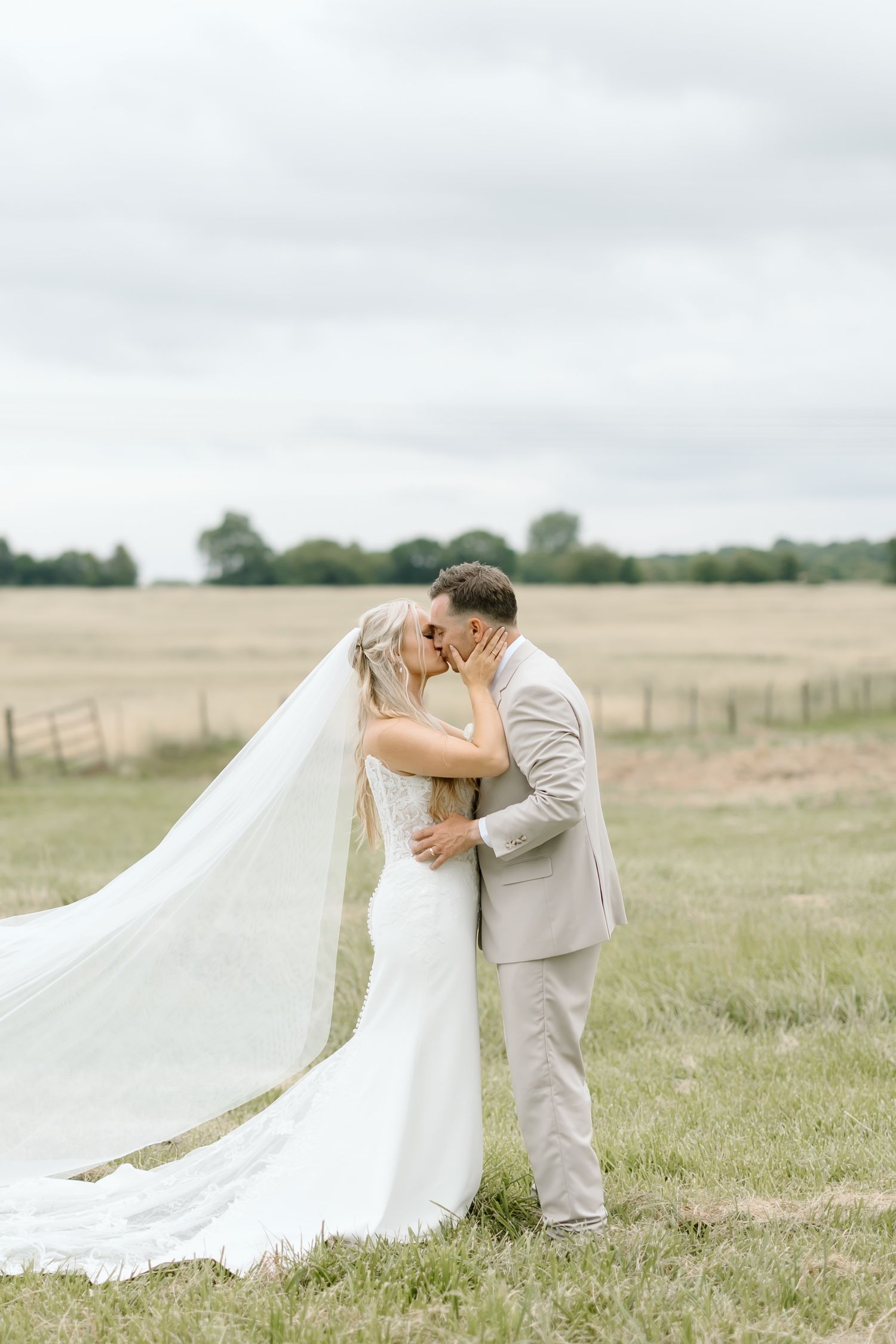 Bride and groom kissing in a field, bride wearing a flowing white gown and long veil on their wedding day.
