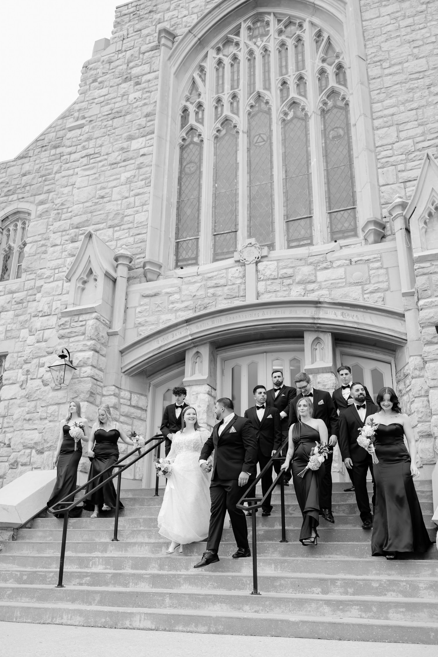 Black and white photo of wedding party descending stone church steps, bride and groom leading groomsmen and bridesmaids.