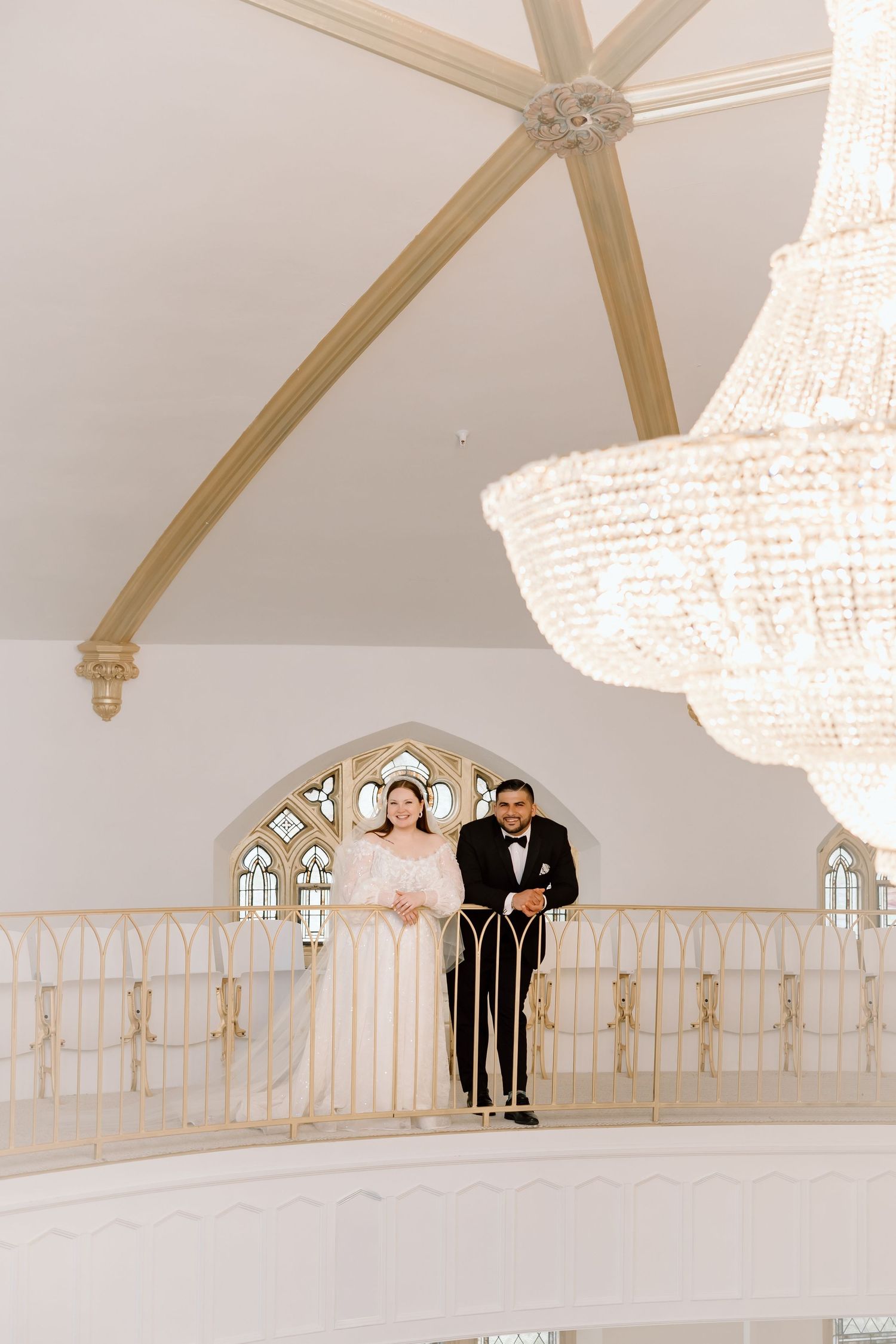 Bride and groom stand on elegant balcony with crystal chandelier in white wedding venue with Gothic arched windows.