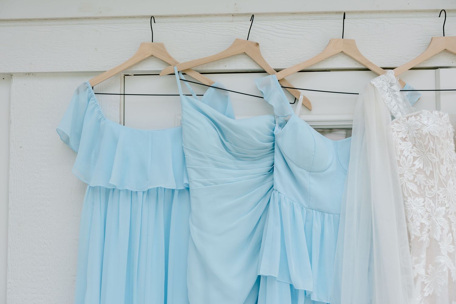 Three light blue bridesmaid dresses on wooden hangers displayed beside a white lace wedding gown.