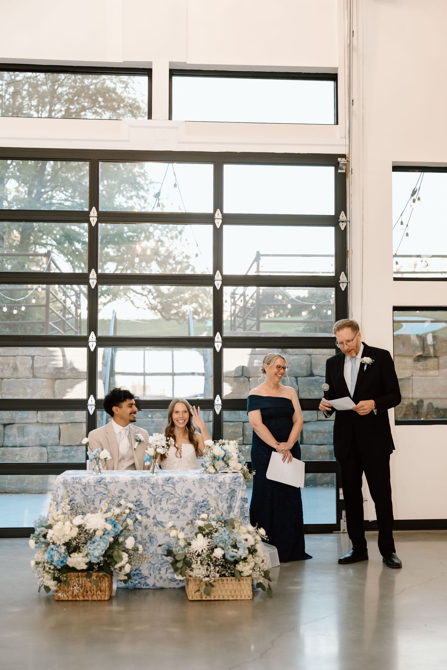 Couple seated at sweetheart table with blue floral arrangements while officiant reads vows in modern venue.