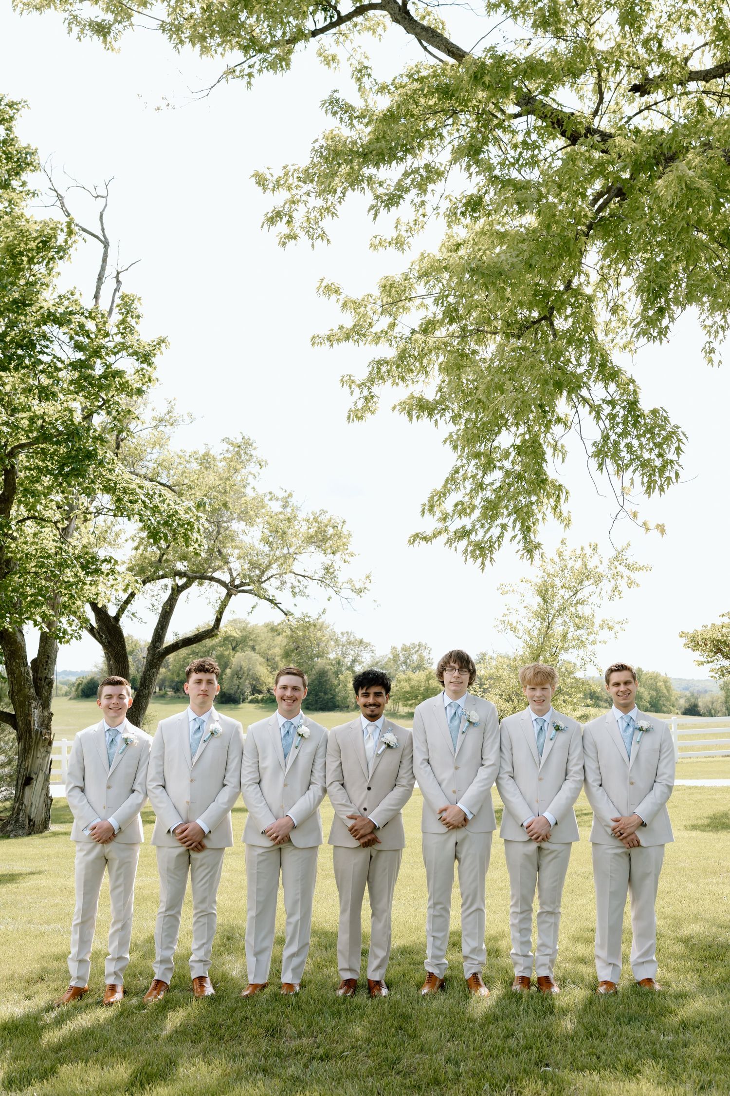 Seven groomsmen in light gray suits with blue ties pose outdoors on a green lawn with trees in the background.