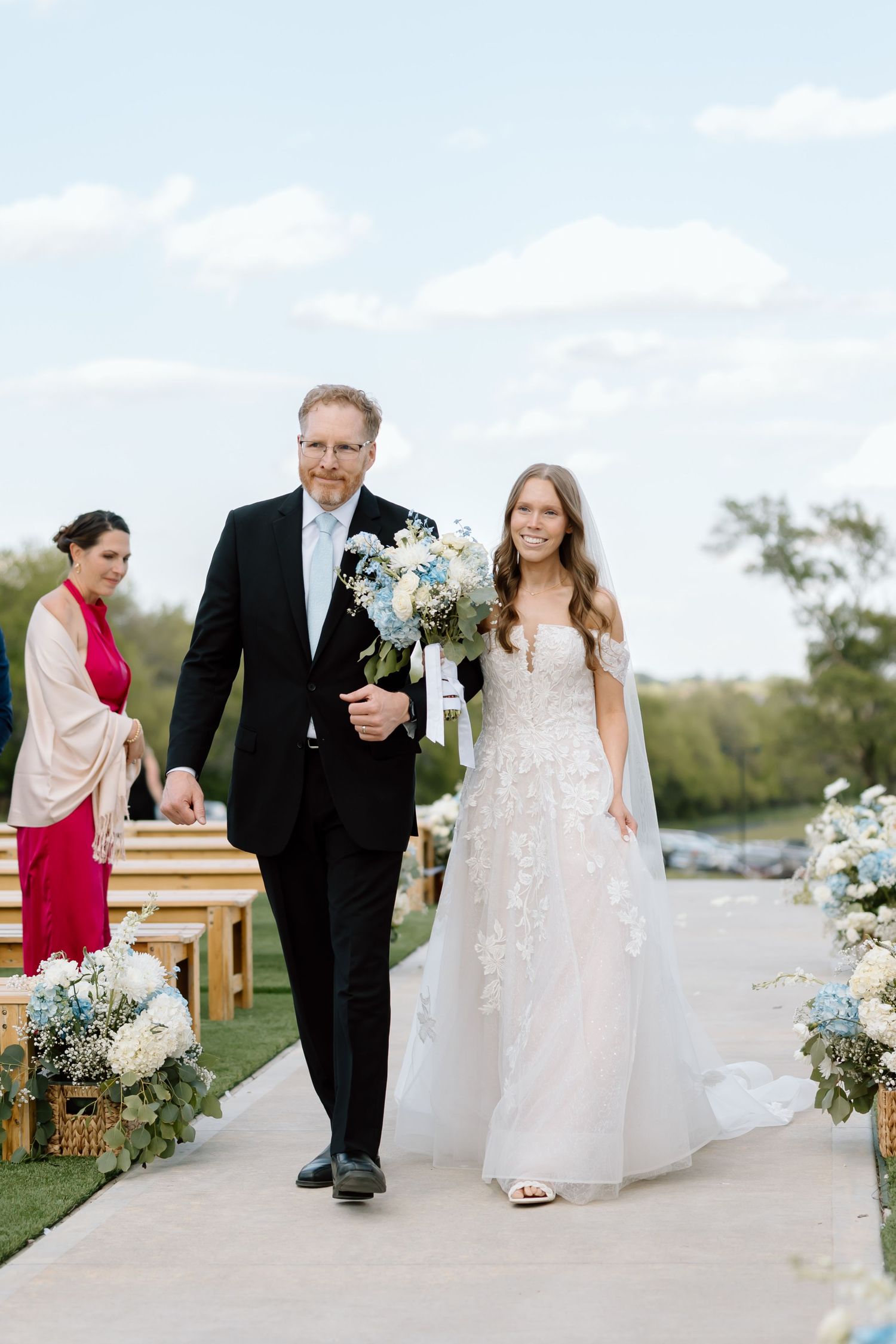 Bride in floral gown walks down outdoor aisle with escort, holding blue and white bouquet at elegant wedding ceremony.