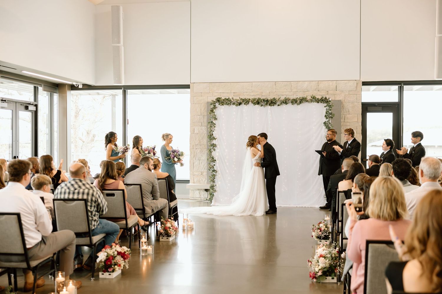 Wedding ceremony in modern venue with floral arch backdrop and seated guests watching couple exchange vows.