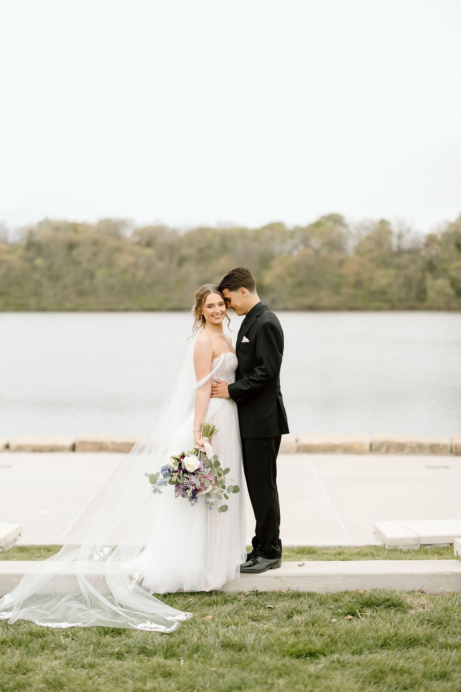 Romantic wedding portrait of couple standing by lakeside with flowing veil.