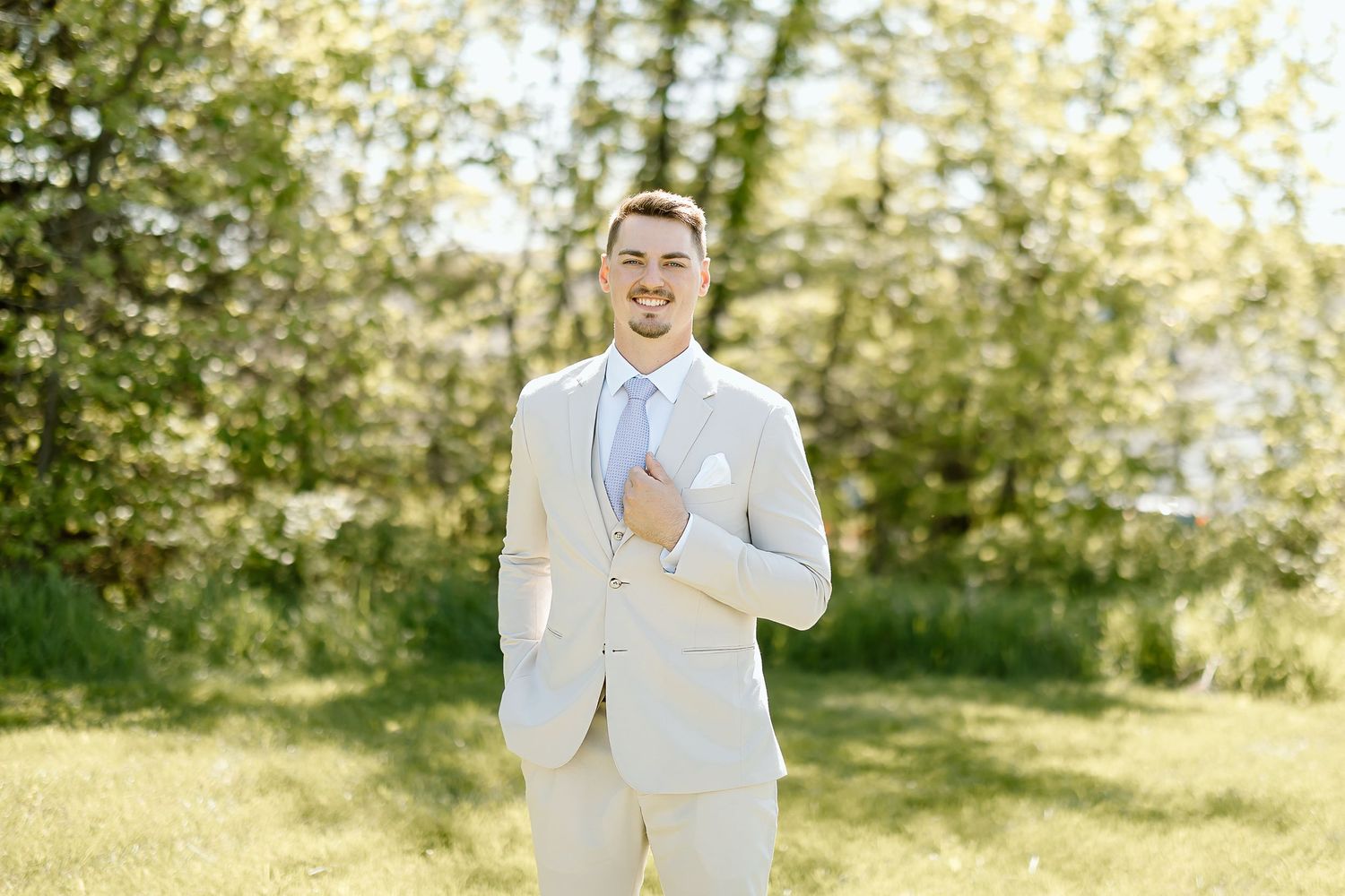 Groom in light suit laughing joyfully in sunny outdoor setting.
