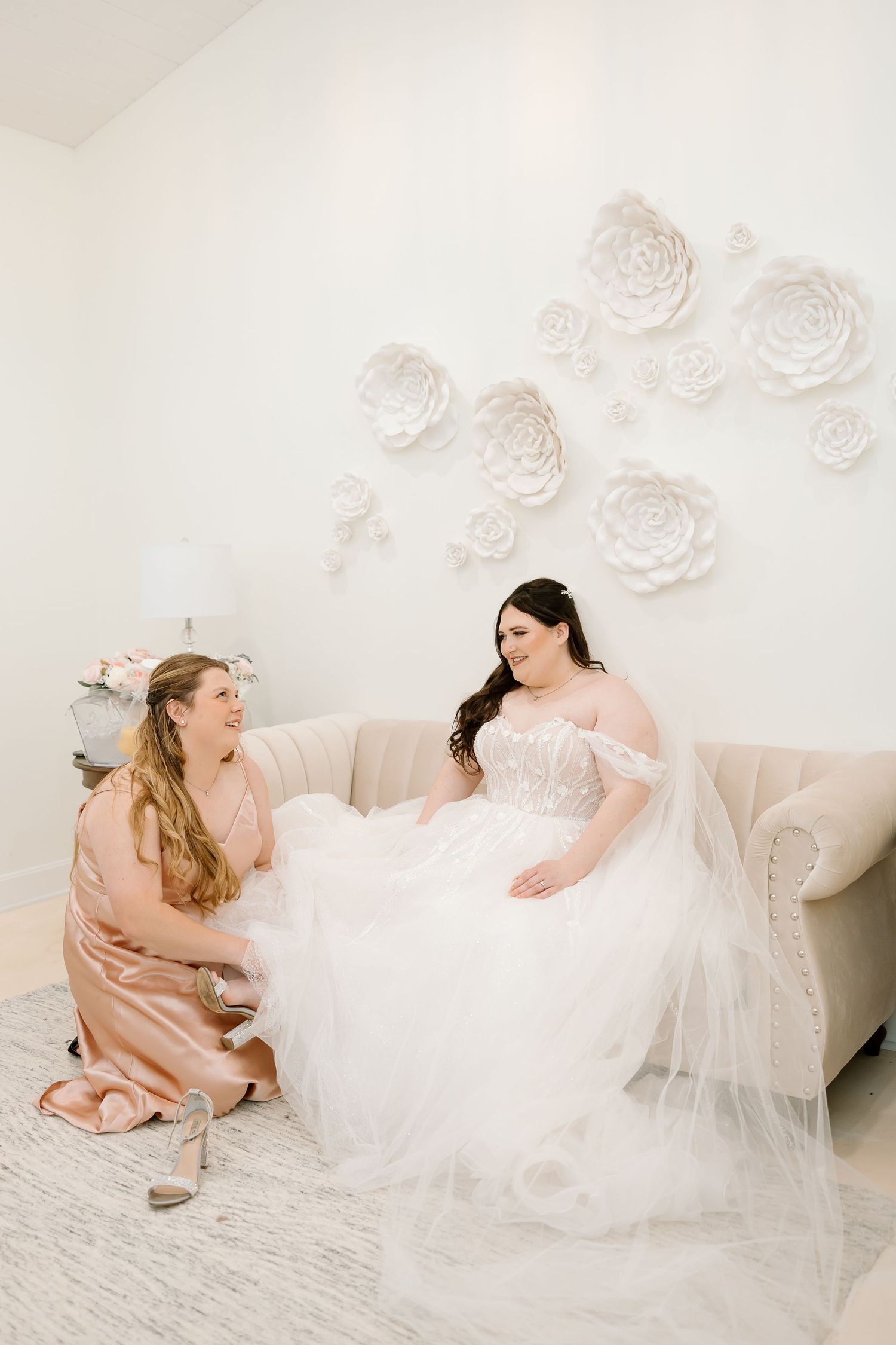 Bride in white gown sits on couch under decorative wall flowers with bridesmaid.