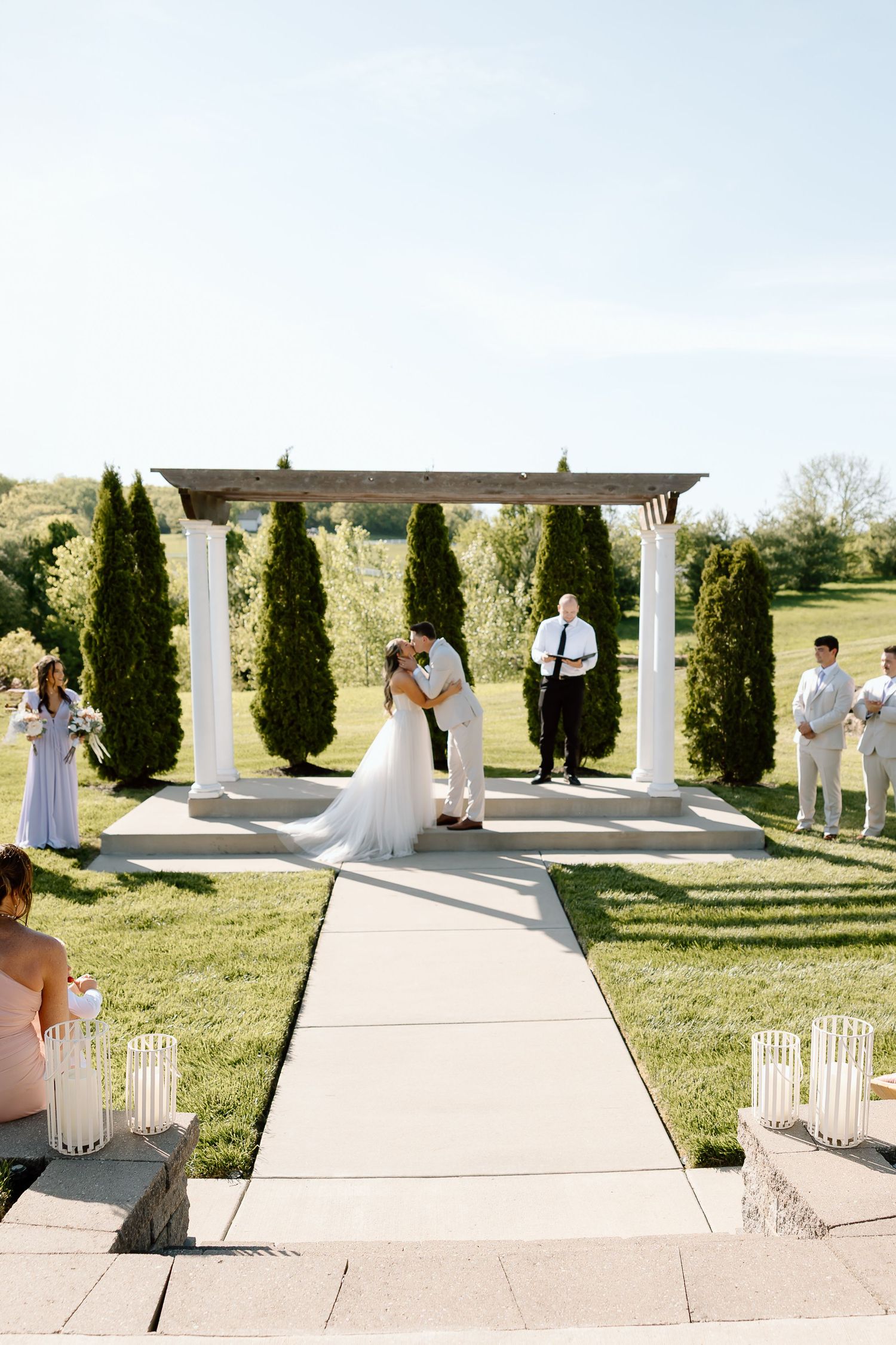Outdoor wedding ceremony under white pergola with cypress trees and lantern-lined aisle.