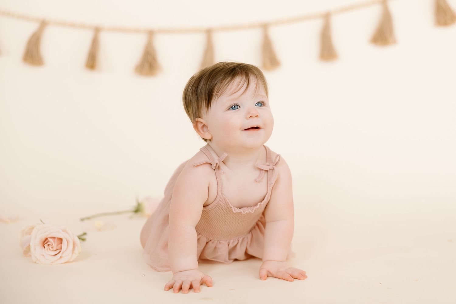 A smiling baby in a pink romper crawls beneath a decorative tassel banner.