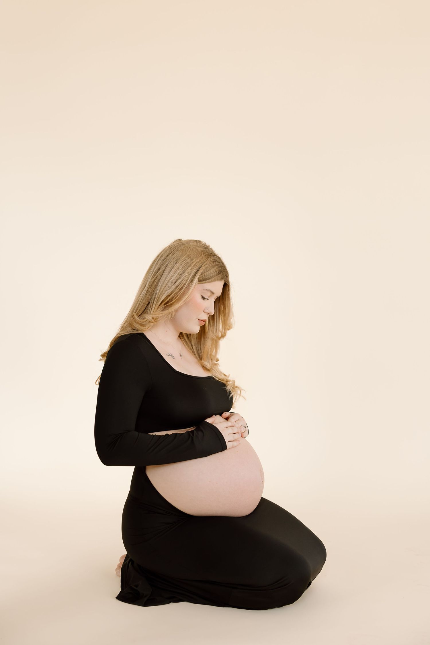 Serene maternity portrait with woman in black dress gazing downward against neutral backdrop.