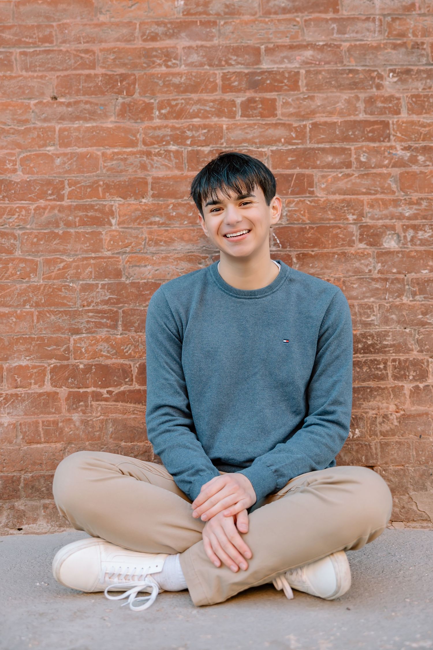 Casual seated portrait against red brick wall wearing blue sweater and khaki pants.