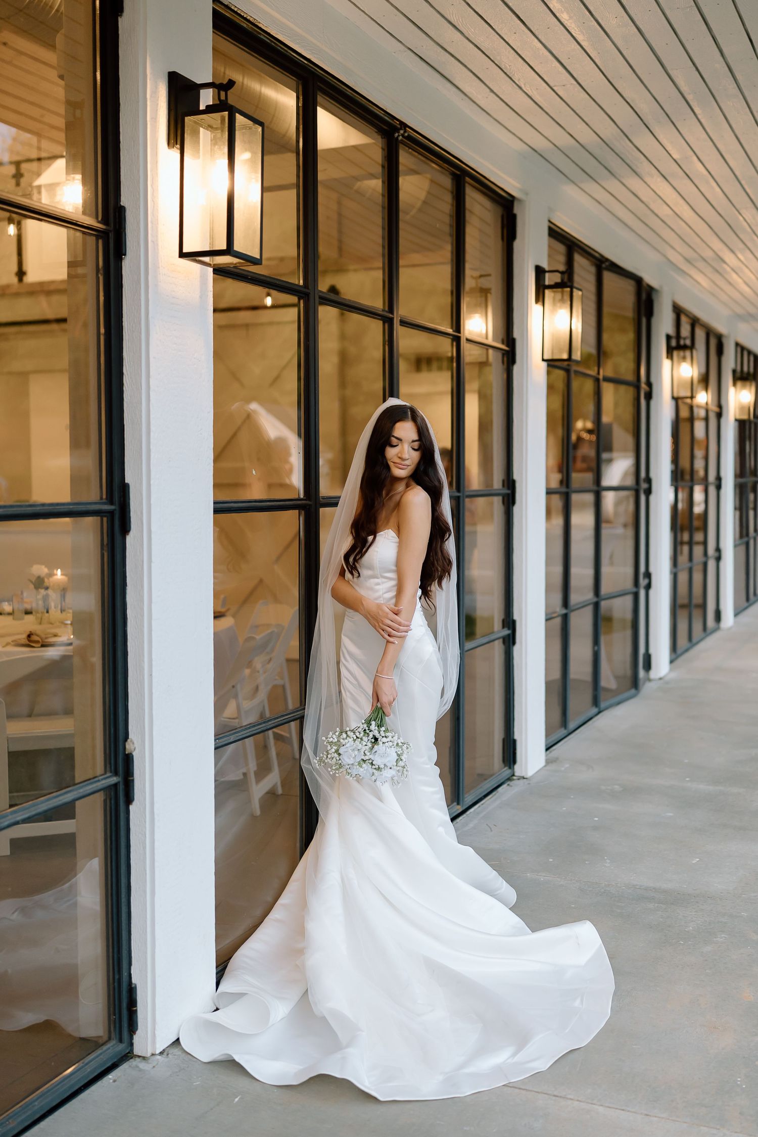 A bride in a flowing white wedding dress poses gracefully in a modern hallway with glass windows and lantern lighting.
