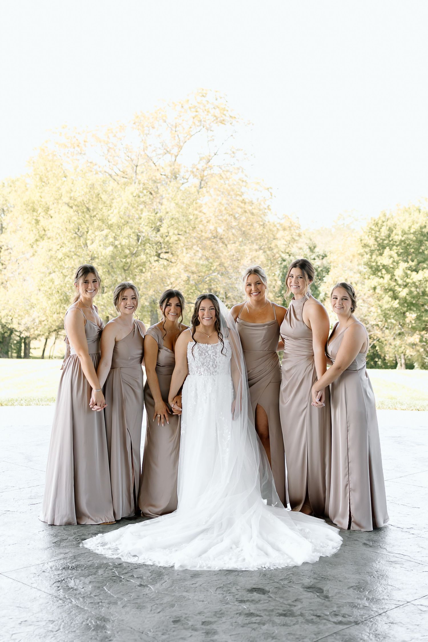 Bridal party in elegant gray dresses and white gown pose together in natural outdoor setting.