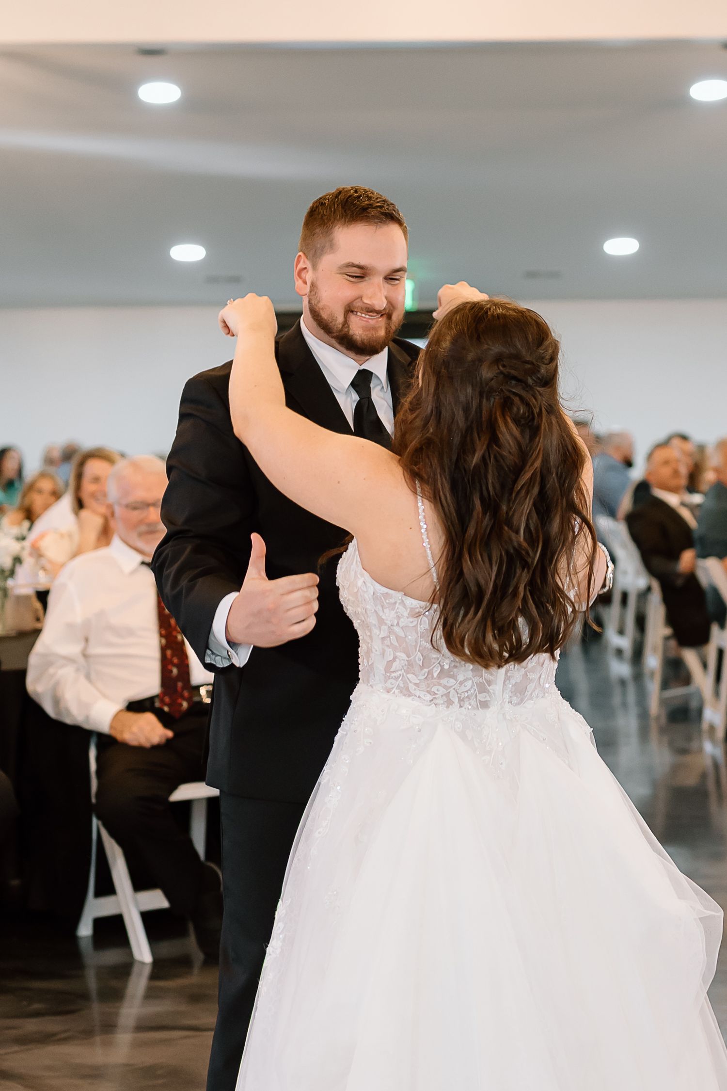 A couple dances together at a wedding reception in a bright white room with guests in the background.
