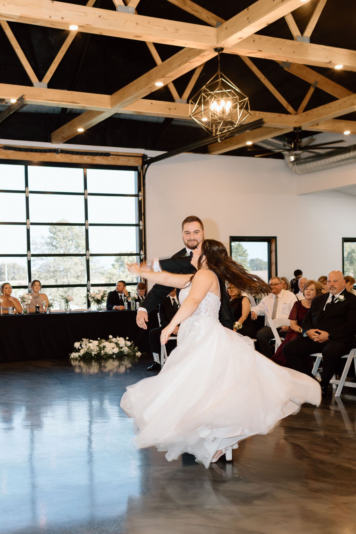 A couple dances their first dance in an industrial-modern venue with exposed wooden beams and geometric lighting fixtures.