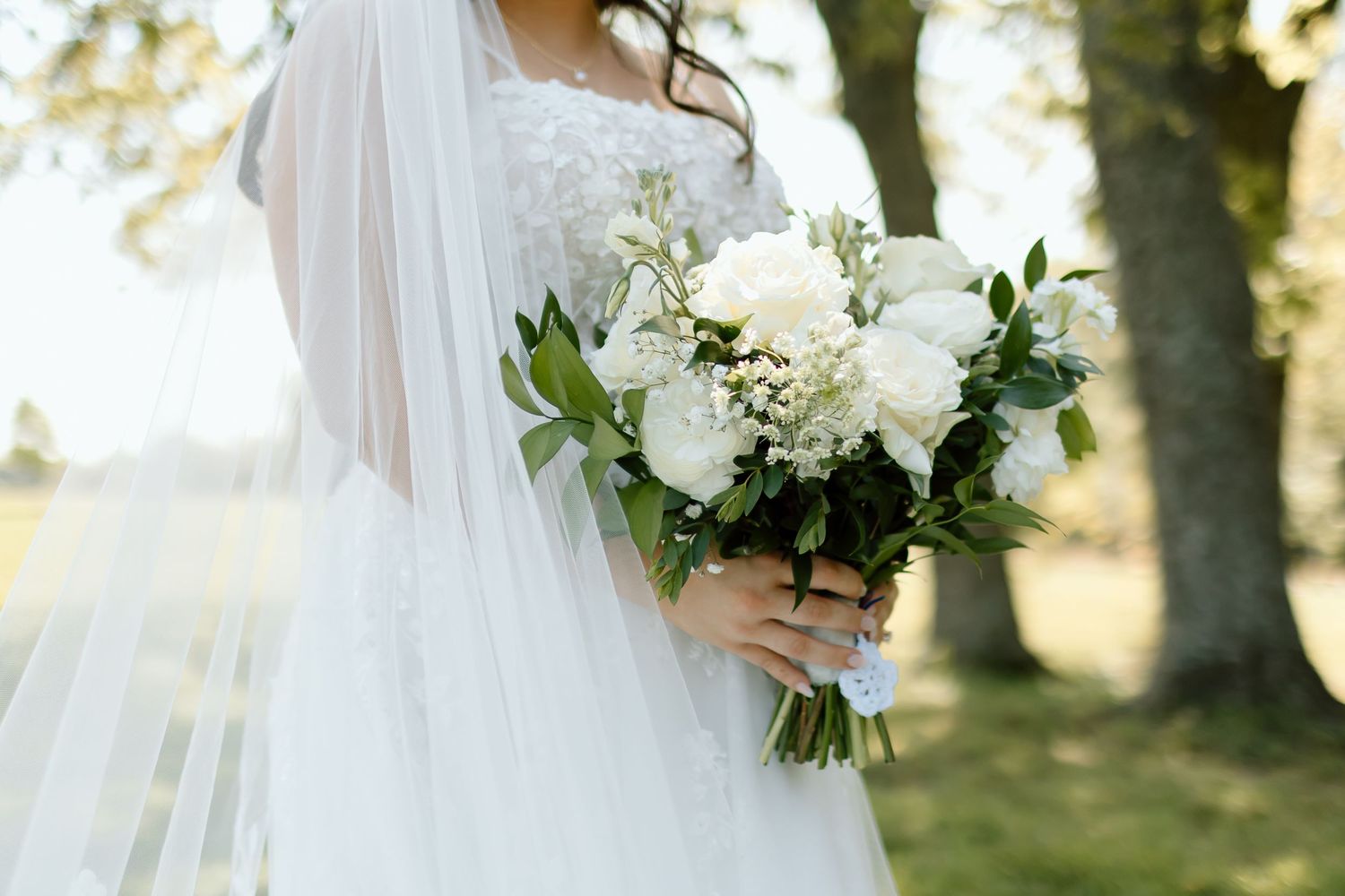 Bride in a flowing white gown holds a lush bouquet of white flowers in a sunlit outdoor garden setting.