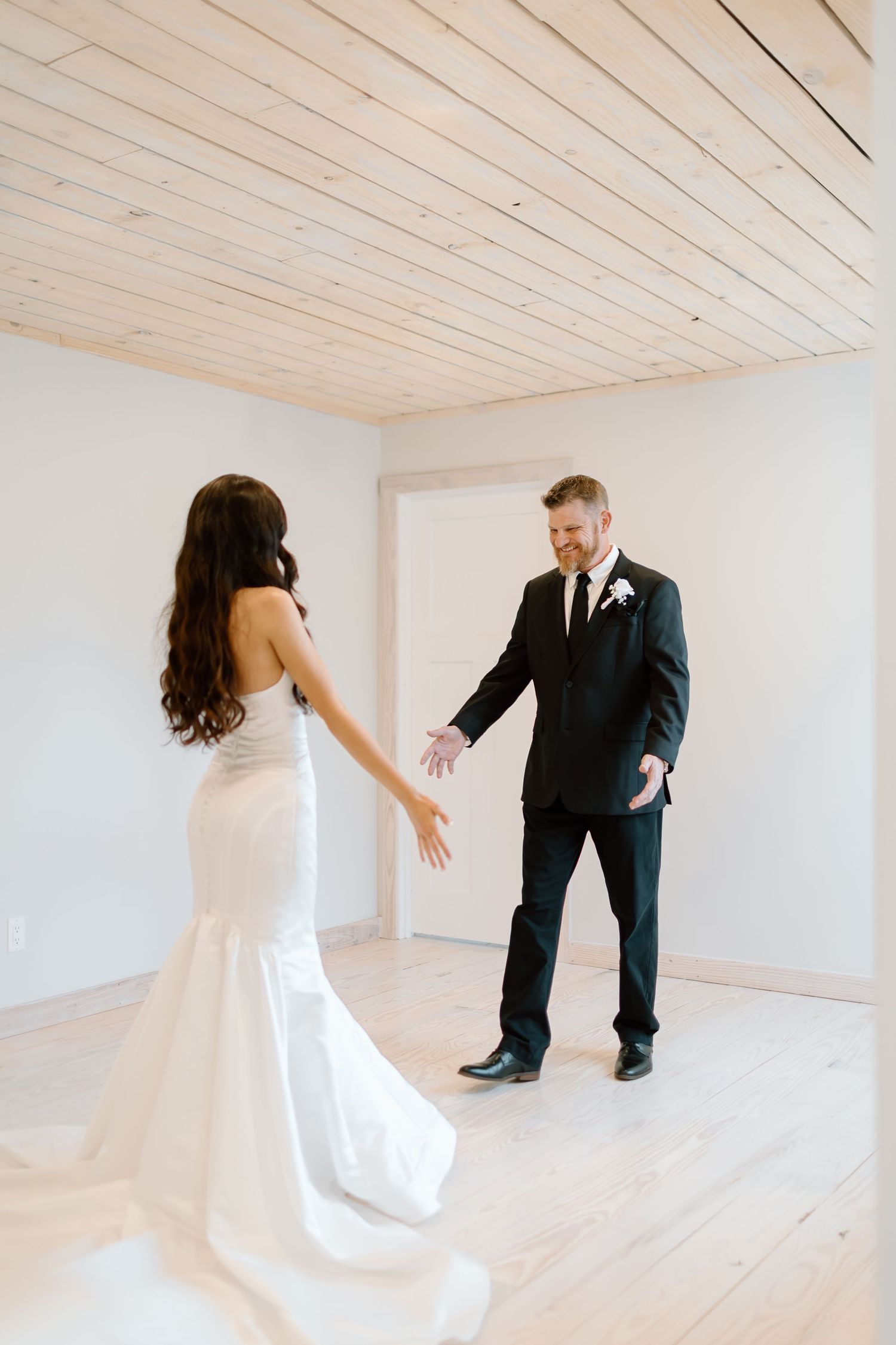 A couple in wedding attire playfully dance and spin in a bright white room with wooden ceiling panels.